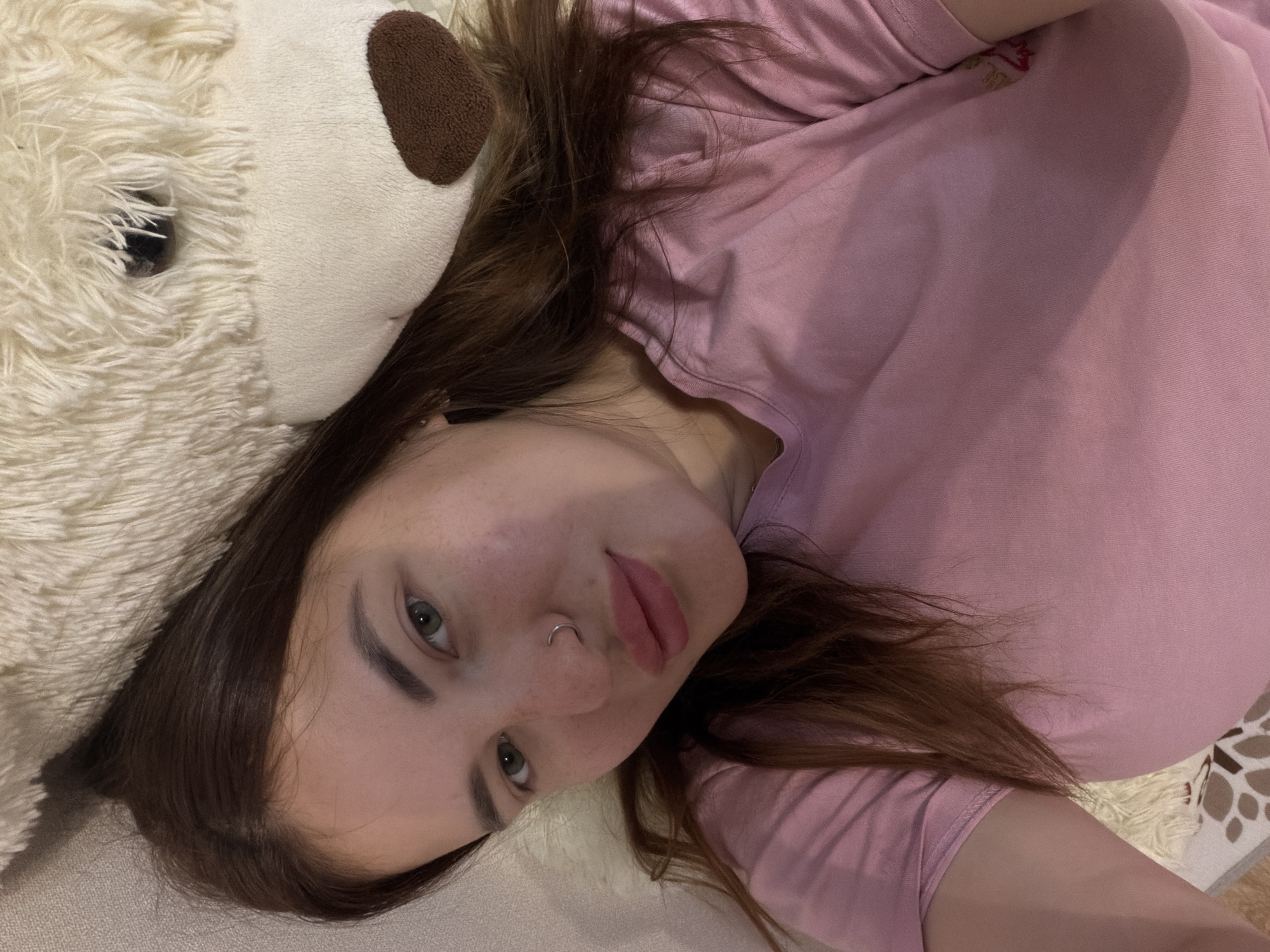 A woman lies on a beige surface, possibly a bed or couch, with a large white teddy bear positioned next to her head. The woman, wearing a pink shirt, has light skin, brown hair, and a nose ring. Her gaze is directed towards the camera. The bear has a brown patch on its head, likely an ear. The environment appears to be indoors, with a patterned design visible in the lower right corner. Location is unknown.