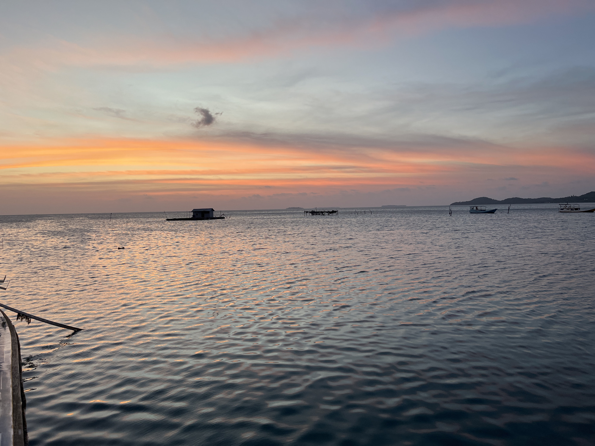 The image captures a serene sunset over the waters of Karimunjawa, Indonesia. The sky is ablaze with hues of orange, pink, and light blue, streaked with wispy clouds. The calm sea reflects the colors of the sky, creating a painterly effect on its surface. In the distance, a small floating structure with a simple roof is visible, likely a dwelling or fishing platform. Several boats are scattered across the water, some appearing to be traditional fishing vessels. Islands or landmasses can be seen on the horizon, silhouetted against the vibrant sky. The foreground shows the edge of a boat or pier, with a wooden plank extending into the water. The overall scene conveys a sense of tranquility and the beauty of nature at dusk.