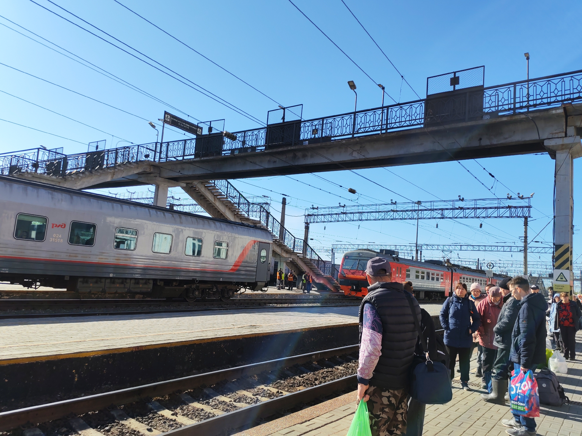 The image shows a train station in Izhevsk, Russia, with a crowd of people waiting on the platform. A gray passenger train is parked on the left track, and a red train is visible on the right. An overhead pedestrian bridge spans the tracks. People are standing with luggage. The sky is blue.