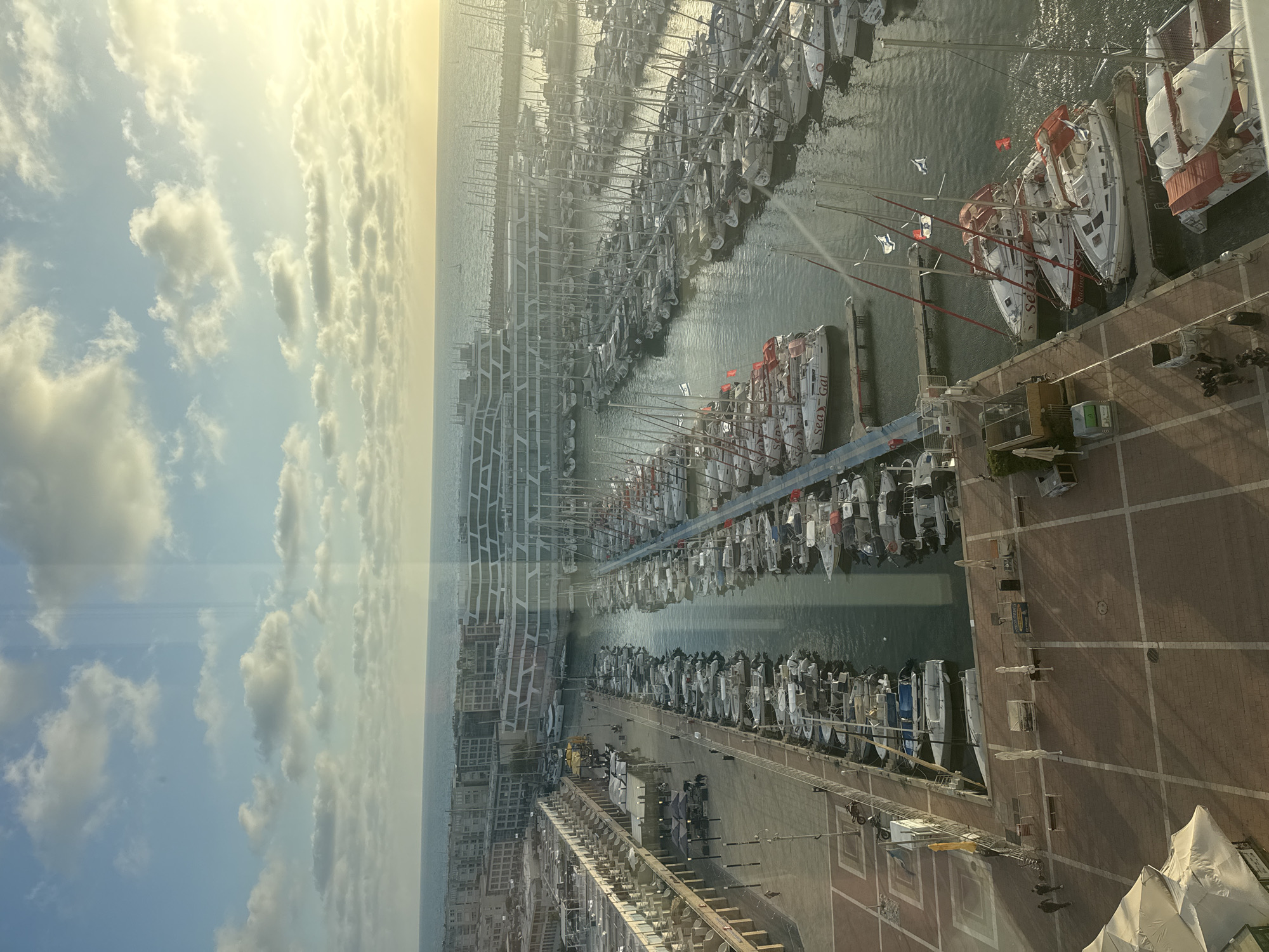 This high-angle shot captures a bustling marina in Herzliya, Israel, under a bright, partly cloudy sky. The image appears to be taken from a tall building or elevated viewpoint, looking down upon the extensive harbor and coastline.
The sky, occupying a significant portion of the image, is a pale blue with scattered white cumulus clouds. A strong glare from the sun is visible in the upper left, indicating a bright, sunny day, likely mid-morning or afternoon.
The central feature is the marina, densely packed with numerous boats, predominantly sailboats, docked in neat rows. Their tall masts create a forest-like appearance against the water. Many boats have their sails furled or covered. The calm water reflects light and the hulls of the vessels. Visible text on some boat hulls includes "Sea-Gal" and partially "Racing". Several Israeli flags are seen flying from the masts of some boats, a clear indicator of the location.
Adjacent to the water is a wide, paved pier or boardwalk, featuring a distinctive brick pattern. Small figures of people can be seen walking along this promenade, adding a sense of scale and activity. Along the landward side of the marina, modern buildings with intricate, geometric architectural designs line the coast. On the pier, there are various temporary structures, including white tent-like covers, as well as scaffolding or ladders, suggesting ongoing work or events. Street furniture, such as signposts and bins, is also scattered across the paved area.
The overall scene conveys a vibrant and active maritime environment, characteristic of a popular coastal destination.