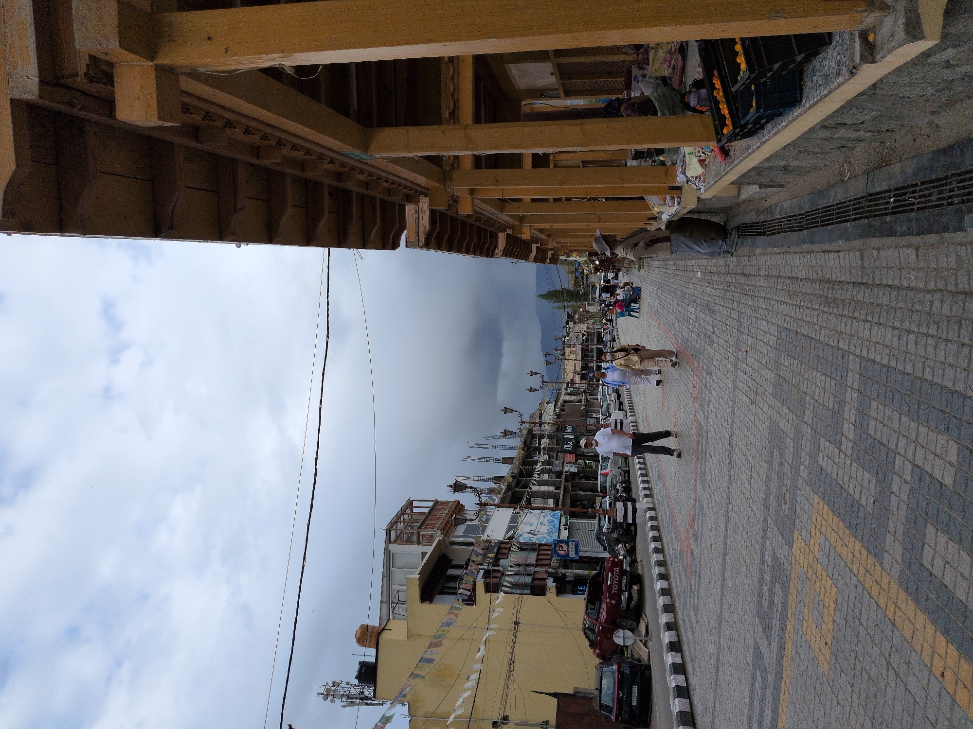 The image depicts a street scene in Leh, India, likely in the morning or afternoon given the bright sky. The perspective is from under a wooden structure, looking down a paved street lined with shops and buildings. Several people are walking along the sidewalk or standing in front of shops. A red Toyota SUV is parked on the side of the road, along with other vehicles. The sky is partially cloudy, suggesting a mix of sun and potential for rain. Colorful prayer flags are strung across some buildings, a common sight in Buddhist regions. The architecture appears to be a mix of traditional and modern, with wooden beams supporting awnings and multi-story buildings. One of the shops has a sign with the word "TOYOTA" visible. The paving stones on the sidewalk are laid in a patterned design, with some yellow accents. The overall atmosphere seems to be one of a bustling market or commercial area.
