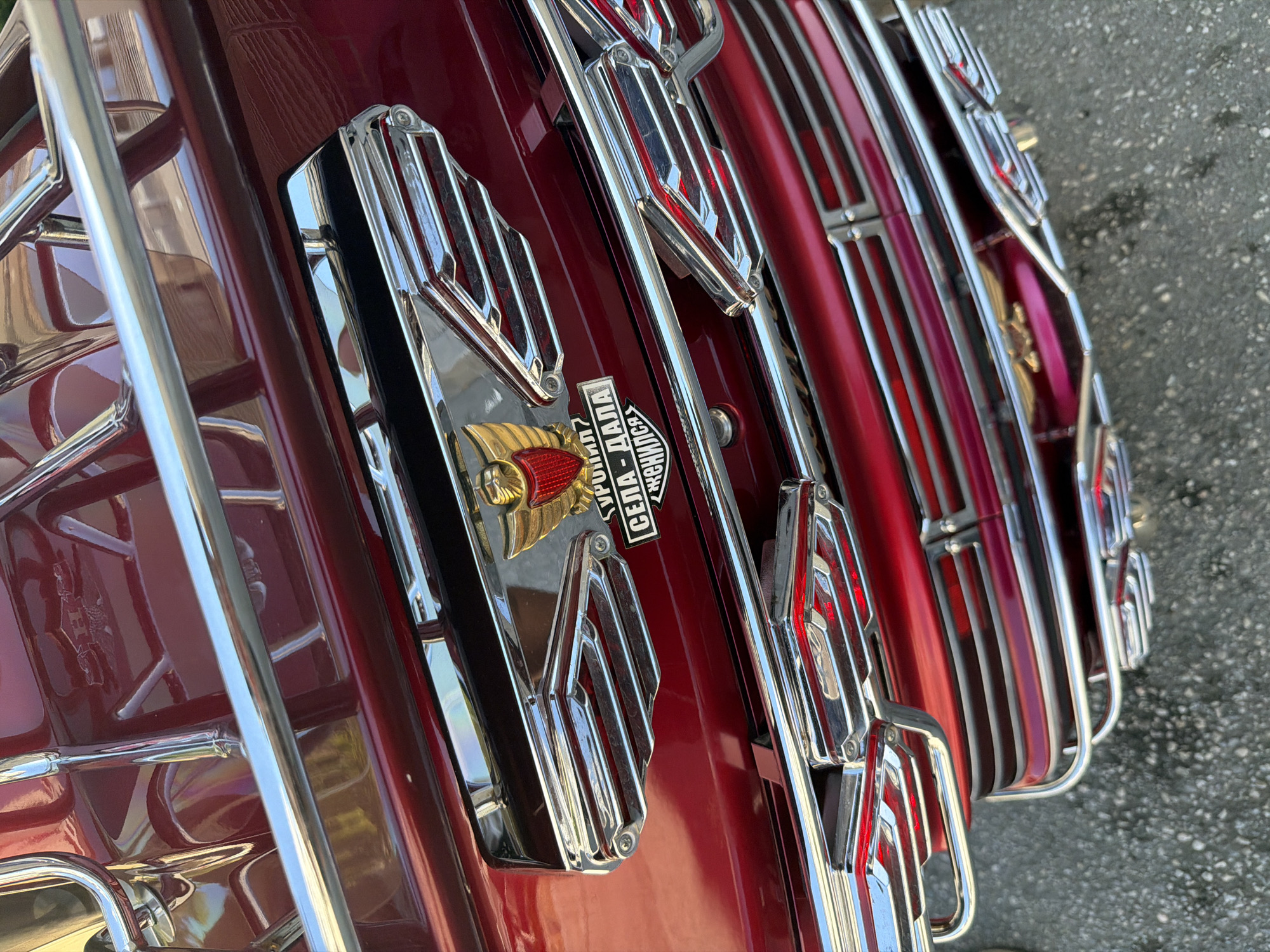 The image shows the rear of a red motorcycle, likely a Gold Wing, adorned with chrome accents and a crest featuring a winged eagle and Cyrillic text. The motorcycle is parked outdoors on a grey asphalt surface. The crest reads "РУПИЋ СЕЛА-ДАЛА СЕНИЛ." In the background, parts of other chrome elements and the motorcycle's red bodywork are visible. The lighting suggests it is daytime, with some reflections on the polished surfaces of the vehicle.