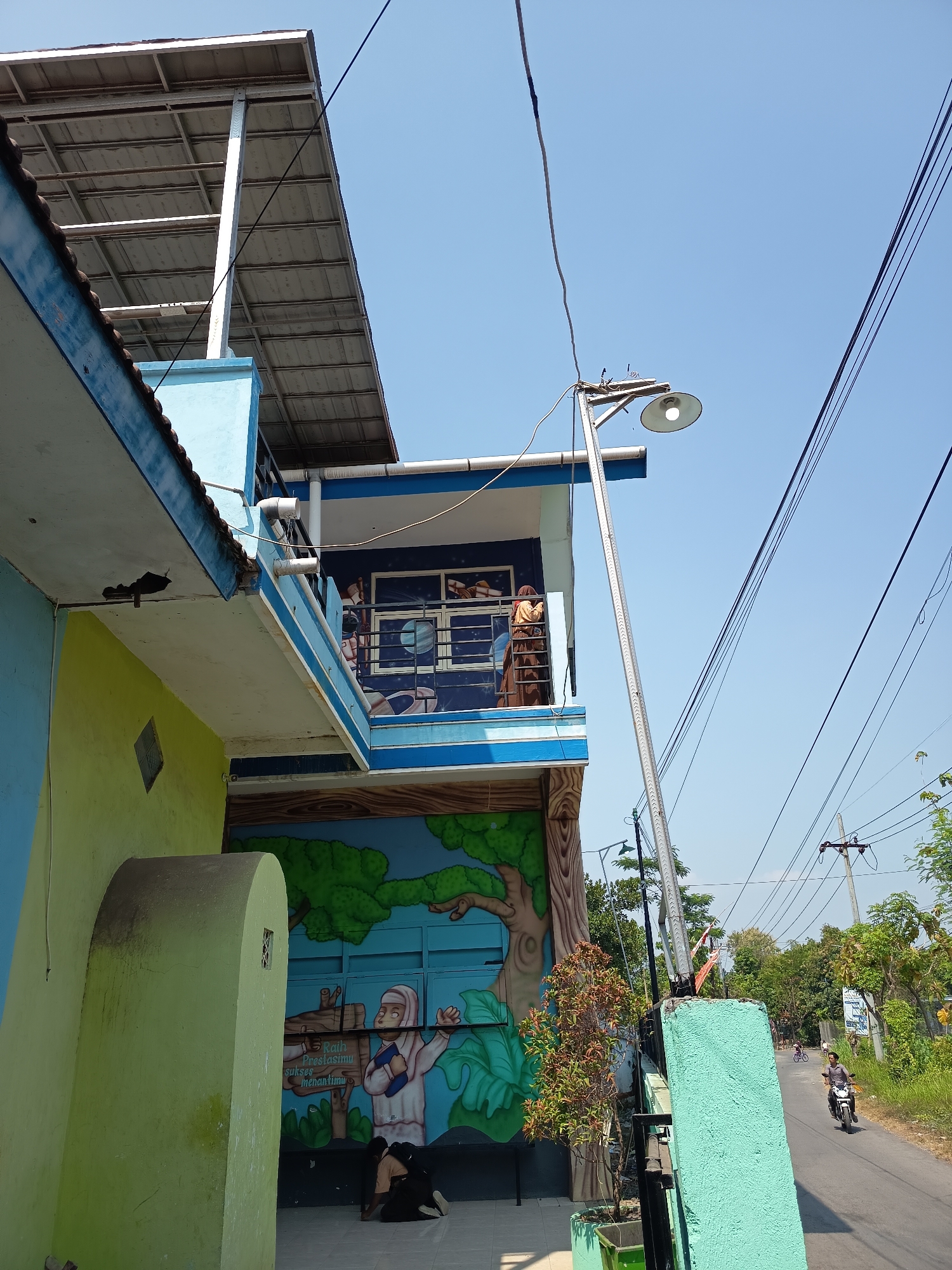 This image captures a scene on a sunny day in Gebangan, Indonesia, with a focus on a two-story building adorned with colorful murals. The building's exterior features a bright lime green wall on the left, transitioning to a light blue upper story with a blue trim. A prominent mural depicting trees, a moon, and a stylized human figure in a hijab holding a book is visible on the lower section of the building, with Indonesian text that translates to "Achieve your success awaits you." Two individuals, one wearing a brown hijab and the other a dark hijab, are partially visible on the second-floor balcony.

To the right of the building, a tall, silver street lamp stands, angled towards the street. A street runs alongside the property, with power lines and poles crisscrossing the clear blue sky. A person on a motorcycle is seen traveling down the road, and other trees and greenery line the street. The overall impression is that of a residential or community area in Indonesia.