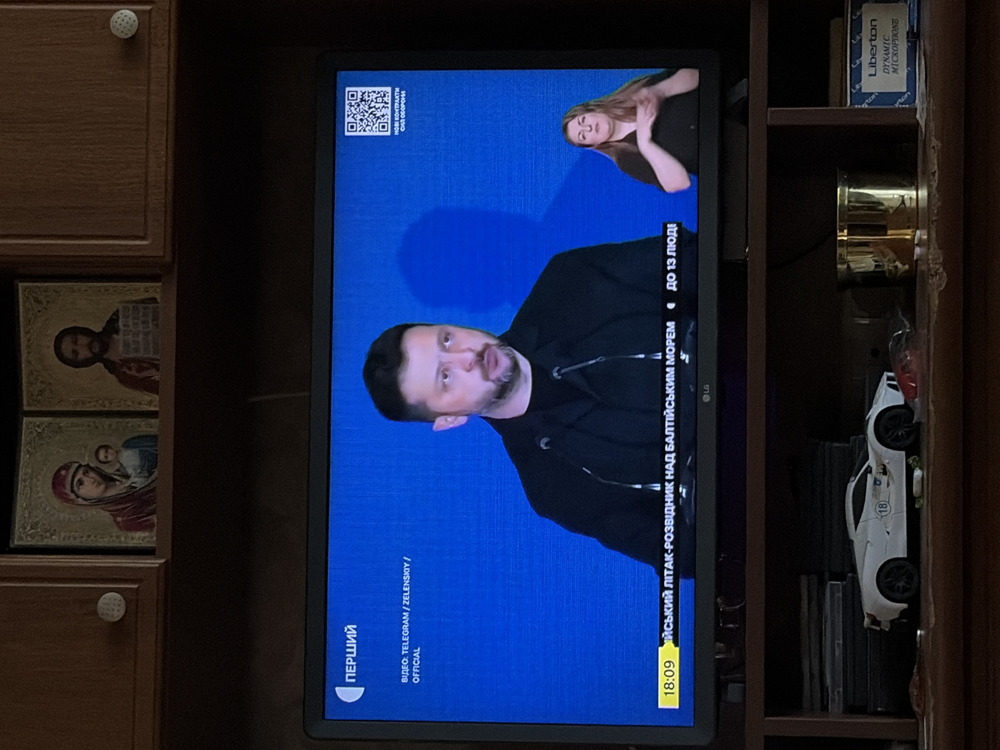 In an indoor setting, a television screen displays a news broadcast. On the screen, a man, identified as Volodymyr Zelenskyy, is speaking, with a sign language interpreter visible to his right. The broadcast's chyron reads "18:09" followed by Ukrainian text. Above the television, religious icons are mounted on a wooden cabinet. Below the television, on a shelf, a white toy sports car, a gold cup, and other objects are visible. The overall scene depicts a home environment with a focus on the television news.
