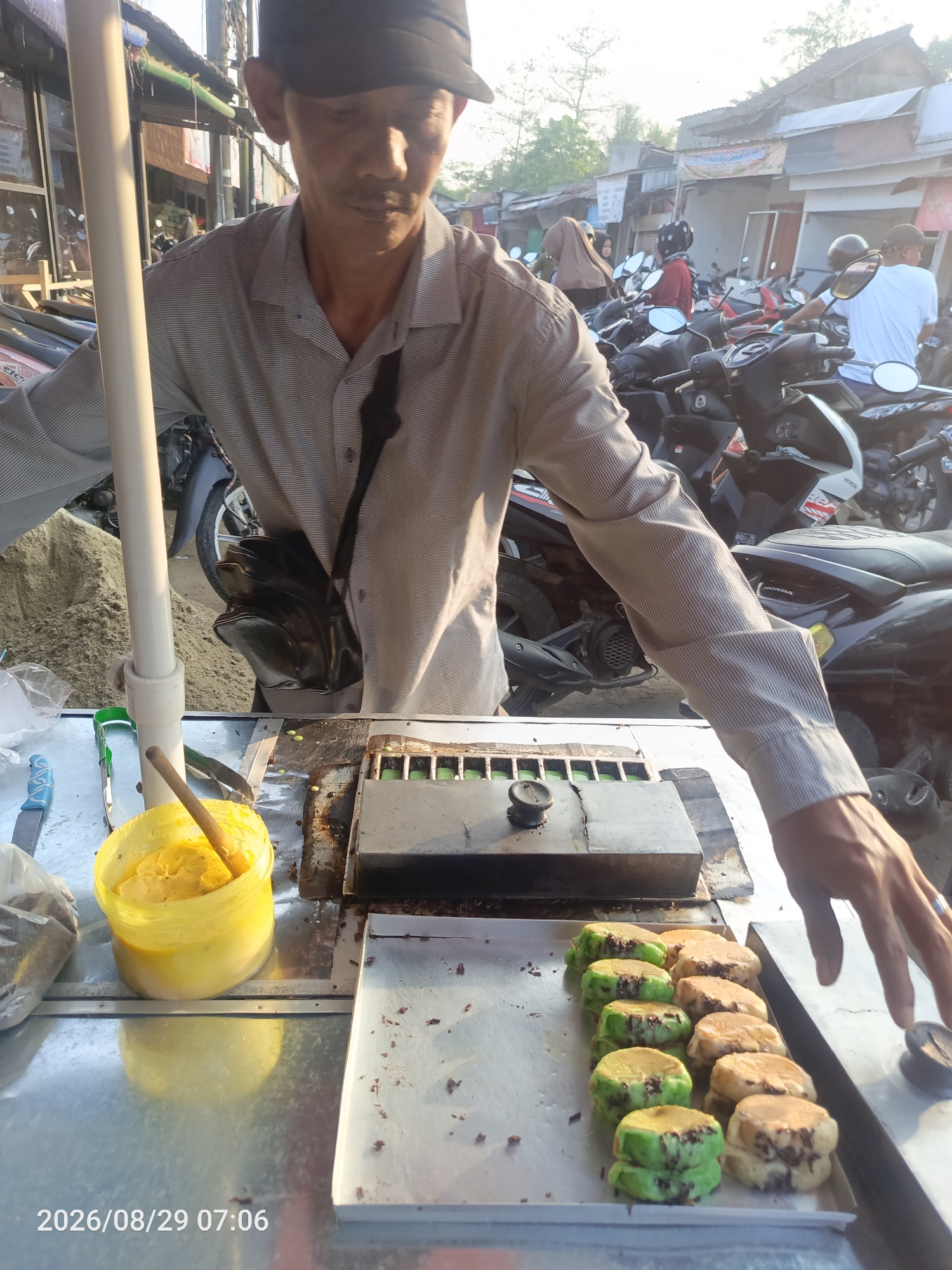 The image shows a street vendor preparing food in Sangiang, Indonesia. A man wearing a grey striped shirt and a dark baseball cap is seen behind a food cart. In front of him, on a metal tray, are several round, flat pastries, some green and some brown, topped with chocolate sprinkles. To the left of the pastries, there is a yellow plastic container filled with a yellow substance, possibly a filling or ingredient, with a wooden spoon sticking out. Behind the vendor, there are many motorcycles parked, suggesting a busy street or marketplace. The background shows buildings with corrugated metal roofs, typical of street-side businesses in Indonesia. The timestamp "2026/08/29 07:06" is visible in the lower left corner of the image.