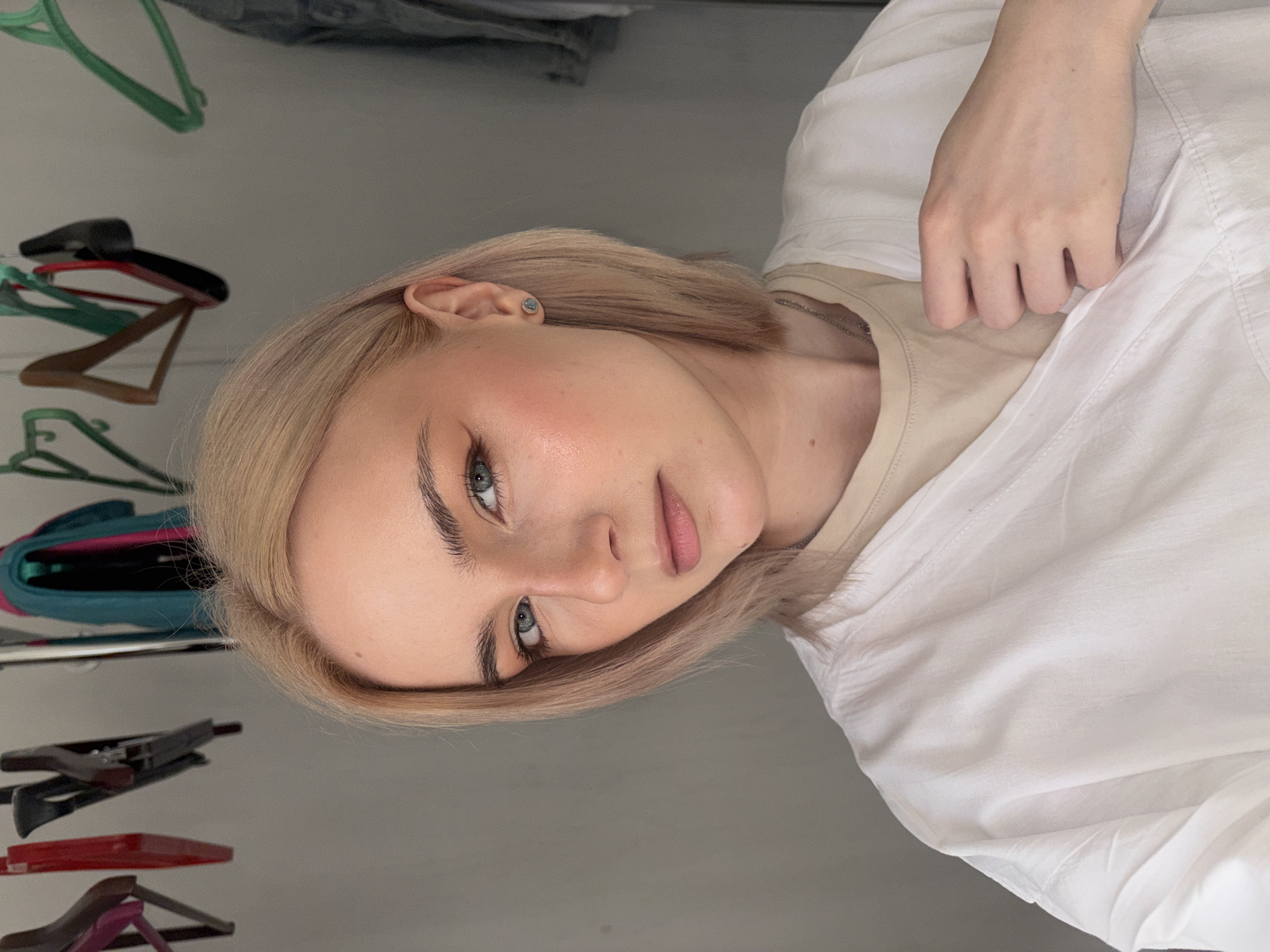 A close-up portrait features a young woman with blonde, chin-length hair and fair skin. She has prominent light blue or grey eyes and subtly made-up features, including defined eyebrows, a soft blush on her cheeks, and light pink lips. She wears a light-colored (possibly beige) undershirt beneath a loose-fitting white shirt or jacket. A small, light blue or green stud earring is visible in her left ear, and a thin chain necklace peeks out from under her collar. Her left hand is casually placed on her white shirt.
The woman is looking directly at the camera with a calm and neutral expression. The background, which is slightly blurred, reveals an indoor setting, likely a personal room or closet. Several clothing hangers of various colors (green, red, black, and brown) are visible on the left side, suggesting a wardrobe or clothes rack. The lighting is soft and even, illuminating her face and creating a gentle atmosphere. There is no visible text or explicit indication of time of day or specific location cues beyond the implied indoor domestic setting.