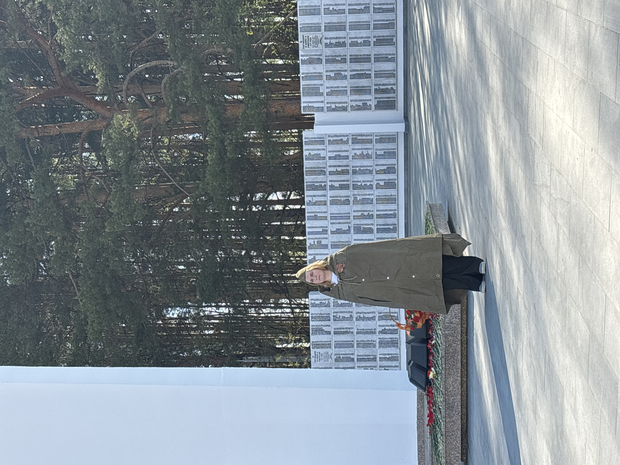 A woman in a dark olive-colored coat and dark pants stands in front of a war memorial. The memorial consists of rows of plaques with names and some kind of graphics. In front of the plaques is a small black, tiered box draped with a floral arrangement including red flowers. The woman is facing the viewer and appears to be looking slightly downward with her right hand raised to her right temple. Trees are visible behind the war memorial. The ground where the viewer is standing is paved with light gray stone blocks. This memorial is located in Chaikovsky, Russia.