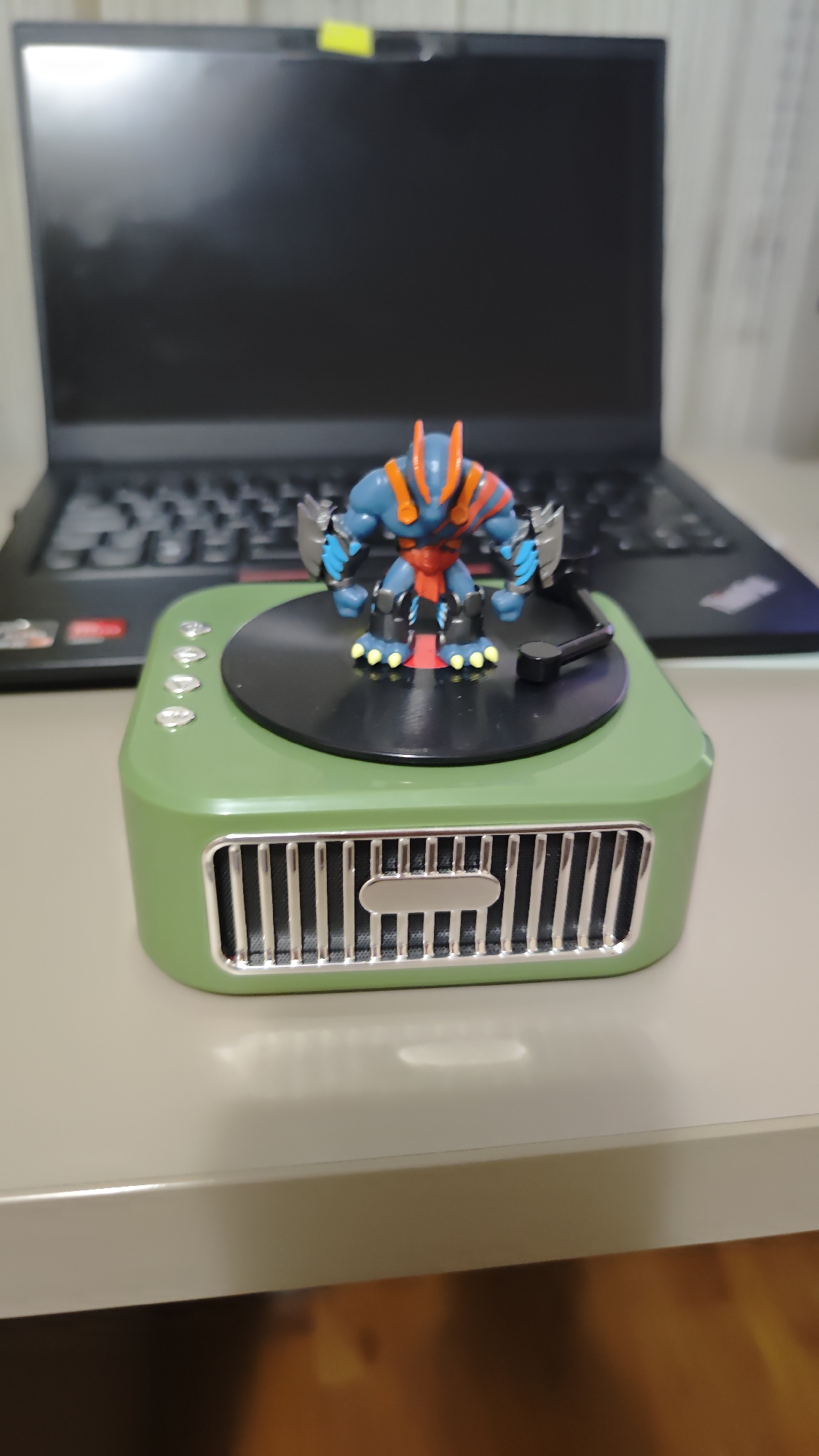 A photograph shows a green retro-style speaker with a black turntable top, on which stands a small action figure. The action figure is primarily blue and red with orange details. A black laptop computer sits behind the speaker, partially out of focus. The scene is set on a light-colored table. Location data is unavailable.