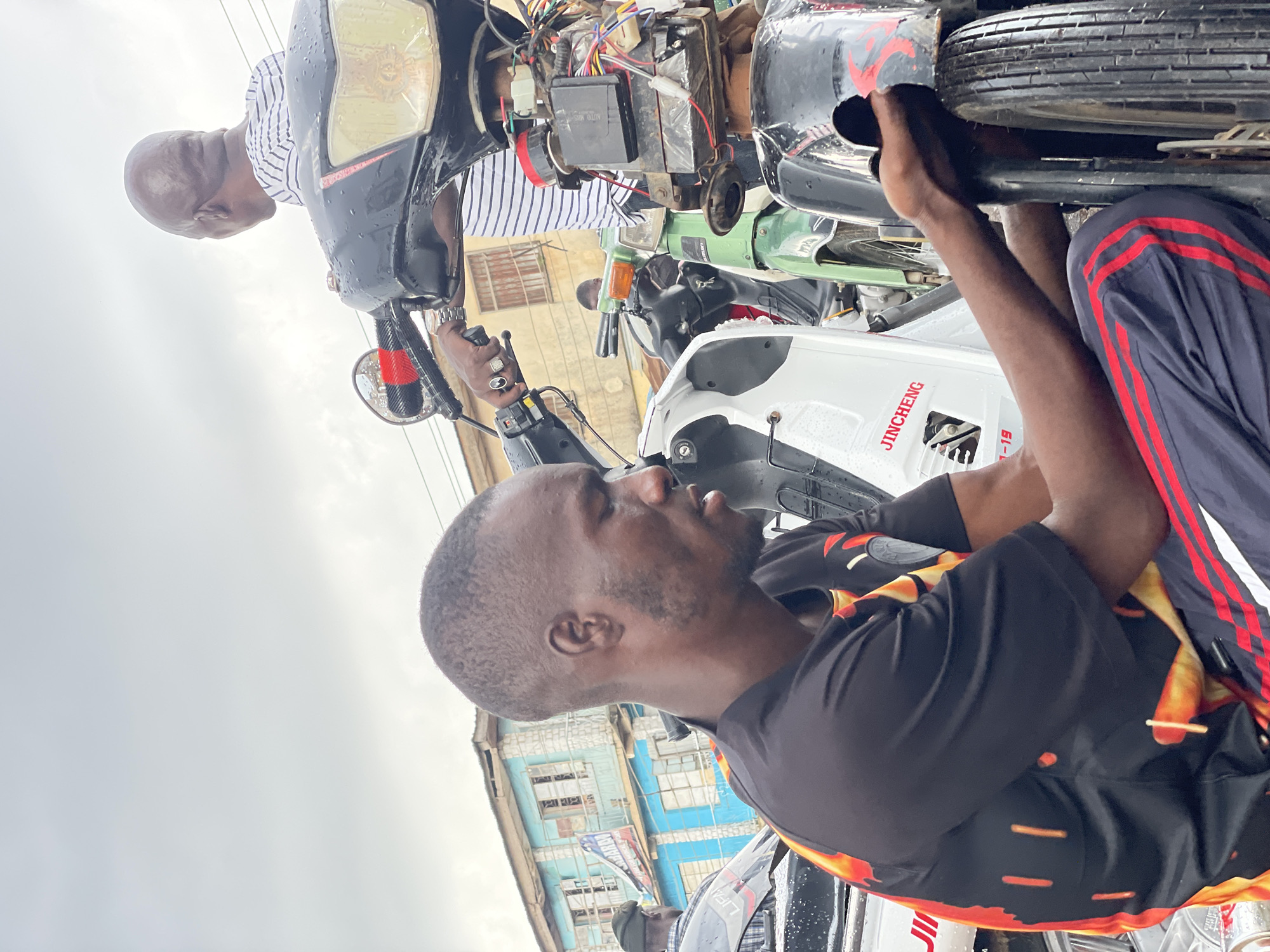 A man is shown from the chest up, his head tilted down and to the right, as he works on a motorcycle in what appears to be an outdoor setting in Jos, Nigeria. He has short hair, dark skin, and is wearing a black t-shirt with a fiery orange and yellow graphic design. His right hand is engaged with the front wheel of the motorcycle, suggesting he is performing repairs. In the background, another man with a shaved head is visible from the shoulders up, wearing a striped t-shirt and looking away from the camera. He is seated on a motorcycle, his hand resting on the handlebars. Several motorcycles are parked close together, with visible wiring and mechanical components on one of them. Buildings with light blue exteriors and visible windows are situated behind the motorcycles. The sky is overcast, suggesting cloudy weather, and the motorcycles appear wet, possibly from recent rain.