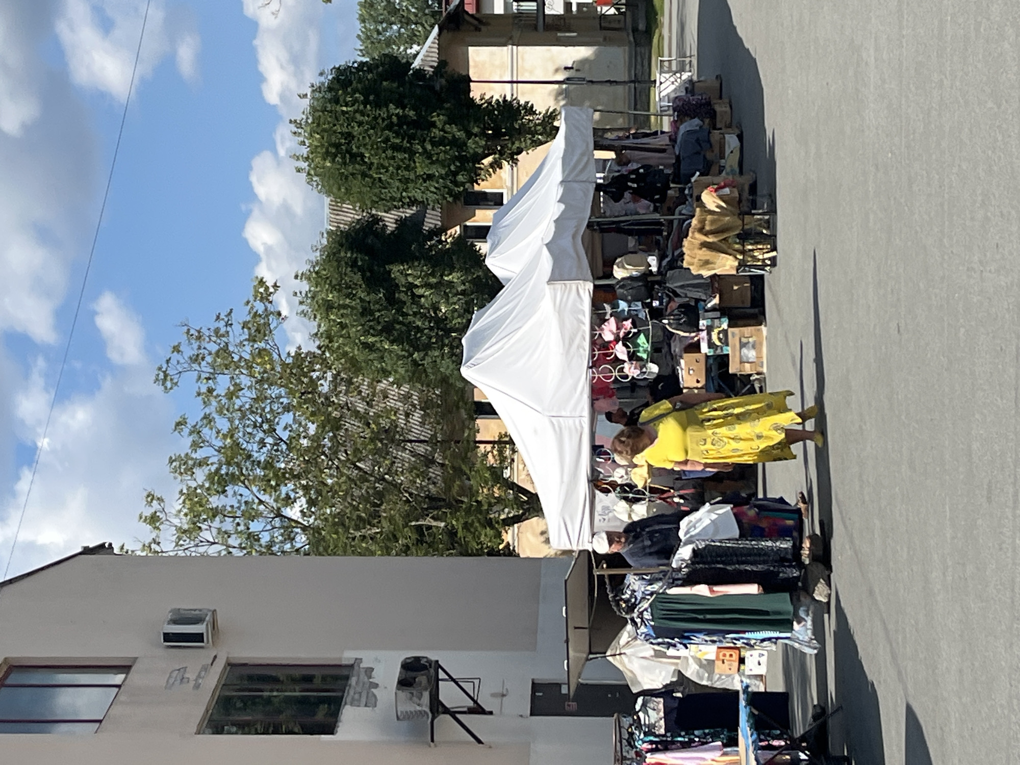 In an outdoor market setting under a partly cloudy sky, a woman in a bright yellow dress walks away from a stall selling clothing. She is the most prominent figure in the foreground. Behind her, a man wearing a white cap and a dark jacket stands near a rack of clothes. The market features two white tents providing shade over various items for sale, including garments, hats, and bags. Clothes are displayed on racks and in stacks on the ground. A building with multiple windows and an air conditioning unit is visible to the left, and trees and more buildings are in the background. The ground is paved asphalt.