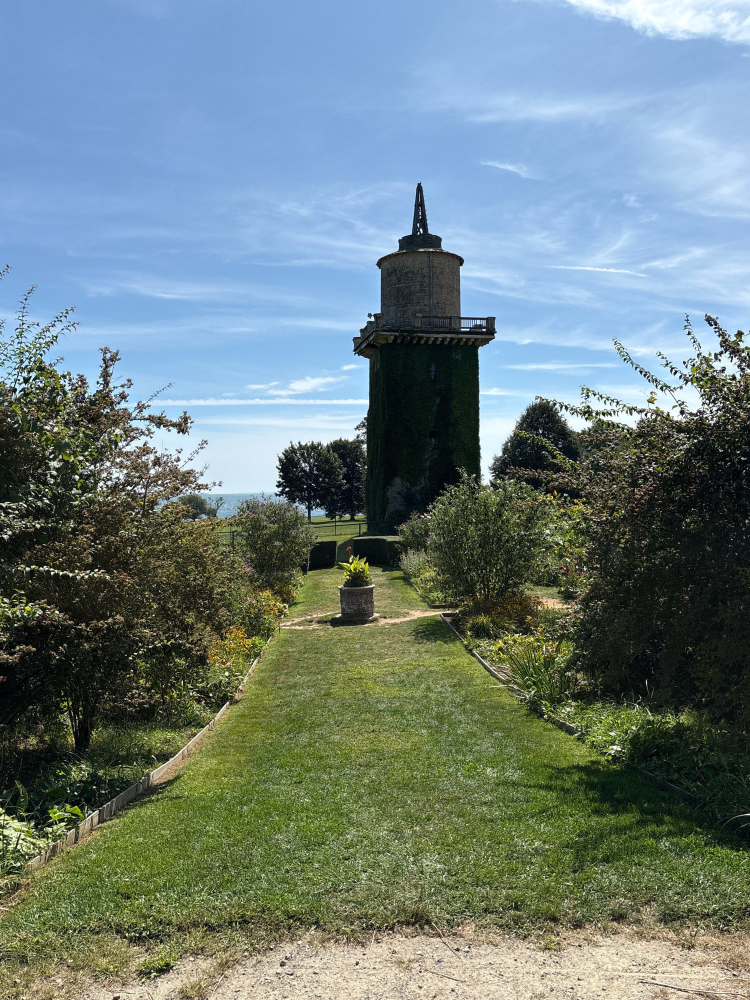 An eye-level, vertical, full shot captures a tall, stone tower covered in ivy and topped with a spire, standing in the center of a lush garden under a clear blue sky with wispy white clouds. A pathway, made of dirt and gravel, leads from the bottom foreground towards the tower, splitting into a well-manicured green lawn that extends up to the tower's base. To the left and right of the path, dense, green foliage and flowering plants fill the garden. The tower has a balcony with a railing encircling its upper section, and a decorative stone planter with a single red-leafed plant sits on the lawn in front of it. In the background, beyond the garden, a body of water, likely the ocean, is visible, with trees dotting the landscape. The overall impression is one of a well-maintained historical site on a sunny day.