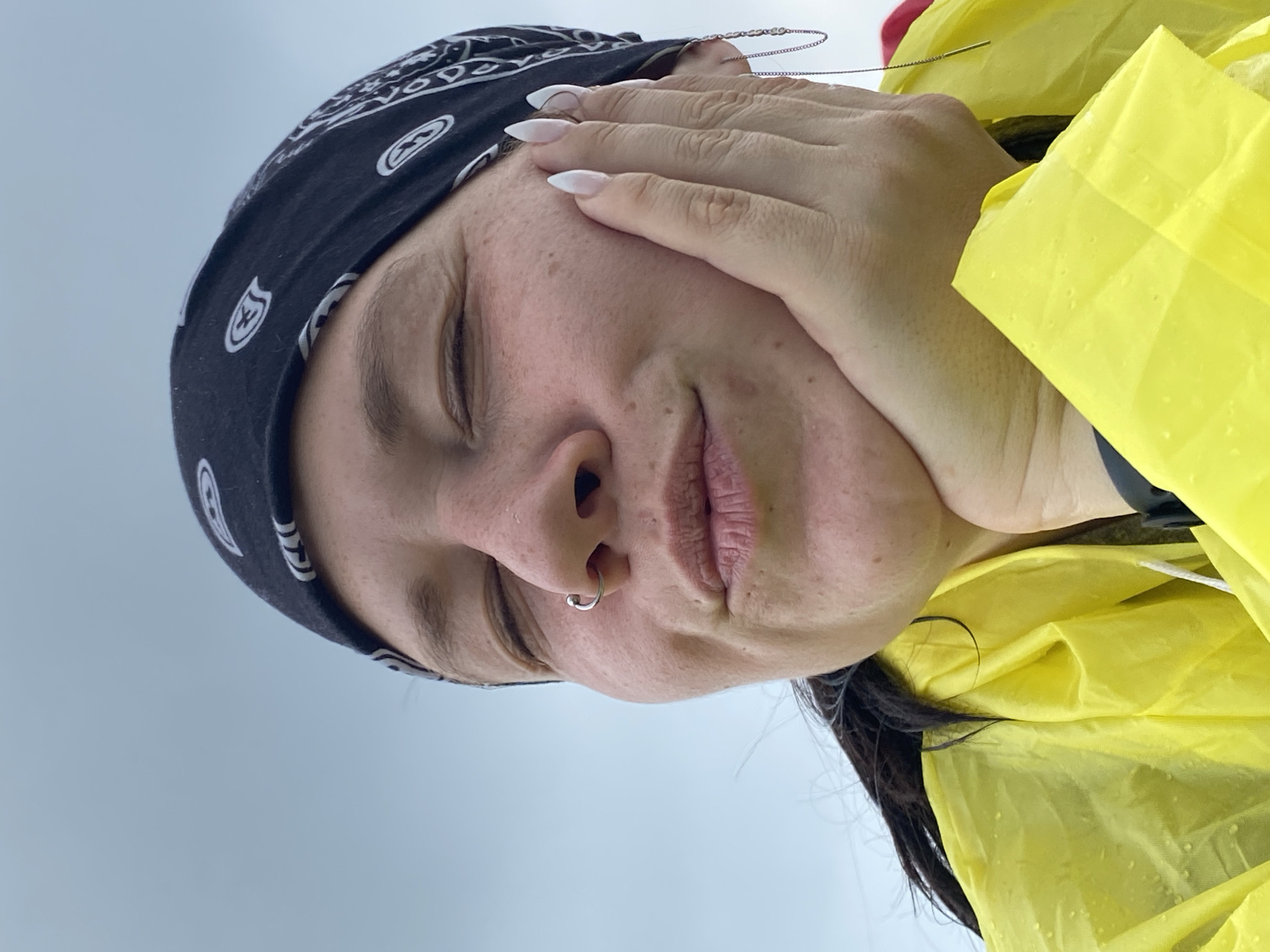 A woman with her eyes closed and lips pursed is resting her cheek on her hand. She is wearing a black bandana with a white paisley pattern, a nose ring, and a yellow raincoat. The background is a light blue sky. The image is a close-up, portrait-style photograph. The scene is not identifiable as being in the city of Рыскулово, Russia.