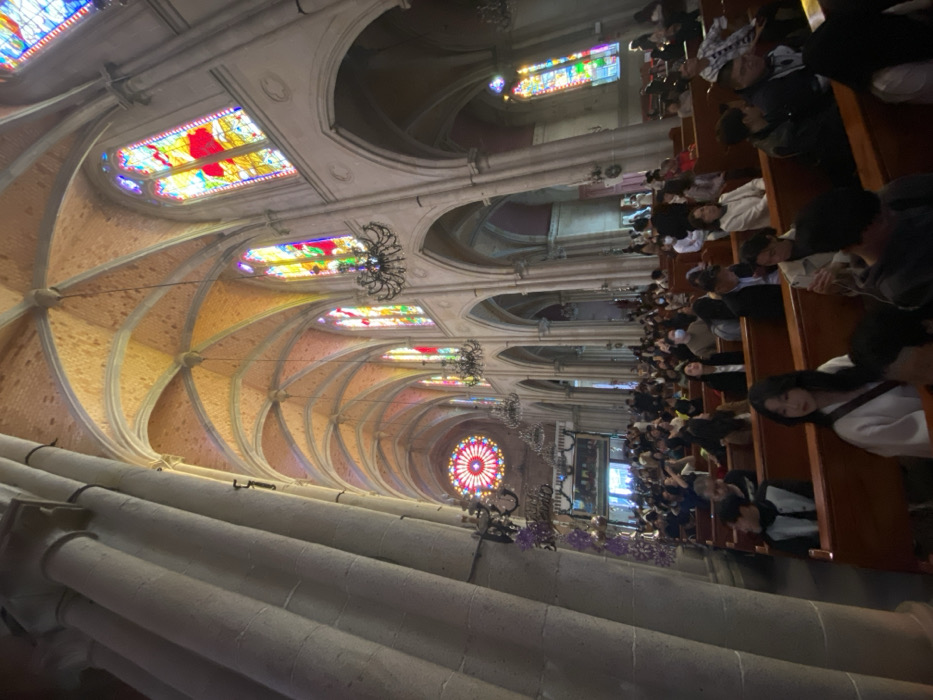 The image displays the interior of a grand architectural space, likely a church or cathedral, captured from a tilted perspective. The photograph is oriented such that the high vaulted ceiling and upper architectural elements are on the left, while rows of pews filled with people are on the right, extending into the depth of the room.
The architecture features elaborate ribbed vaults and arches across the ceiling. Numerous rectangular stained glass windows, vibrant with color, line the upper walls, allowing light to filter into the space. Towards the far end of the hall, a large, intricate circular rose window is prominently visible. Several ornate black chandeliers hang from the ceiling, providing additional illumination. Large, pale stone columns with fluted details line the nave.
On the right side of the image, dark wooden pews are densely packed with a large congregation of people. They are seated, appearing attentive and observant, facing towards the front of the hall (where the rose window is located). Individuals are dressed in varied attire, from casual to smart casual. Some purple floral arrangements are visible in the foreground, possibly adorning a railing or a front section of the pews.
The scene suggests a daytime event, with natural light illuminating the interior through the stained glass. The atmosphere appears to be one of quiet assembly or a service. This location is contextually identified as Guangzhou City, China. There is no visible text within the image.