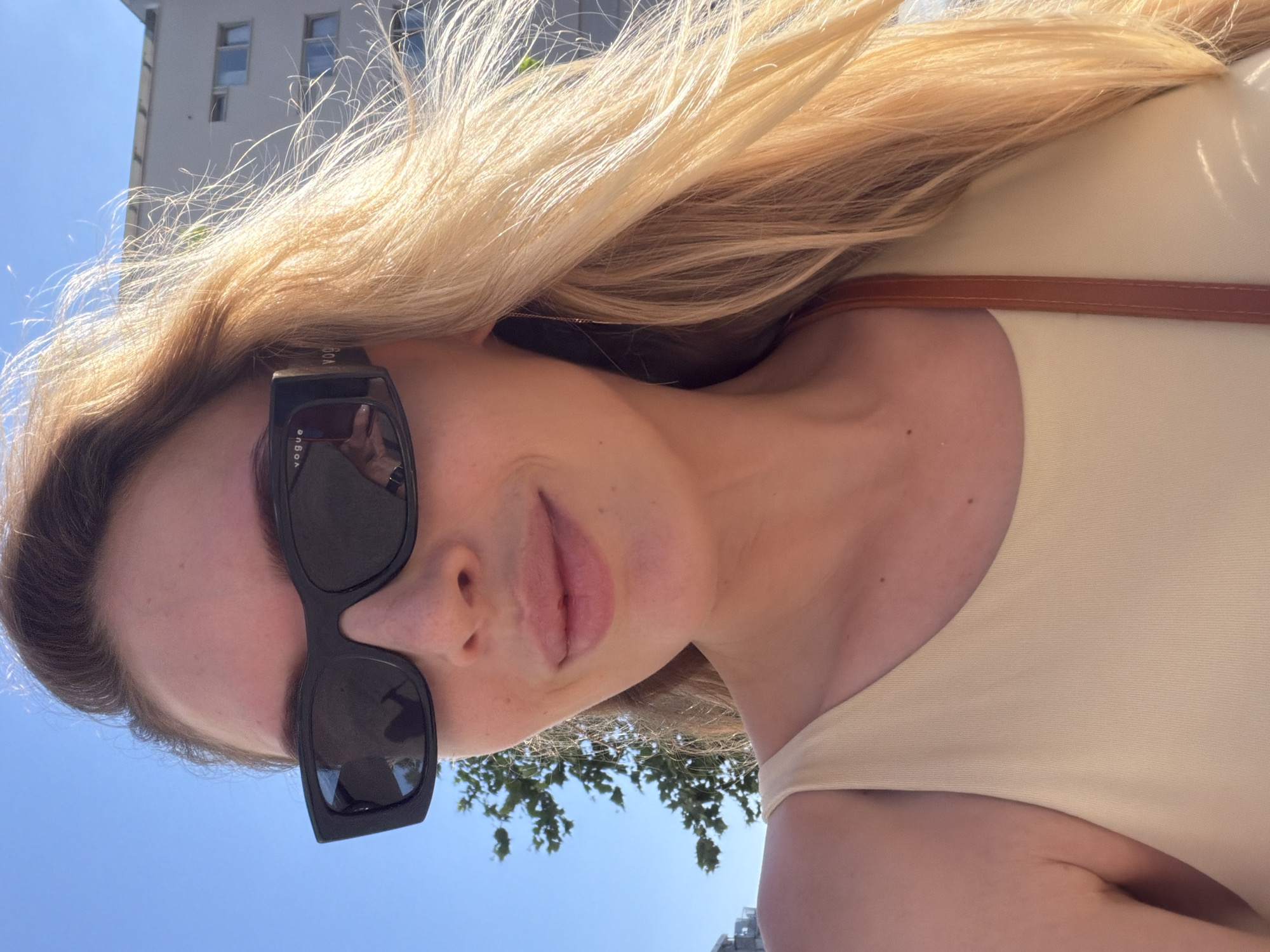An outdoor, close-up, selfie-style photograph shows a woman with blonde hair and fair skin. She is wearing large, black, rectangular sunglasses with the brand name "VOGUE" visible on the side. The sunglasses reflect a person holding a phone, indicating the selfie was taken with a phone. She is wearing a light beige tank top and has a thin brown strap visible over her right shoulder, suggesting a bag or garment. Her lips are slightly parted, and she appears to be smiling. The background includes a clear blue sky, a green tree with leaves, and a modern building with windows. The lighting suggests it is daytime. The context of the city of Budva, Montenegro, is not explicitly visible in the image.