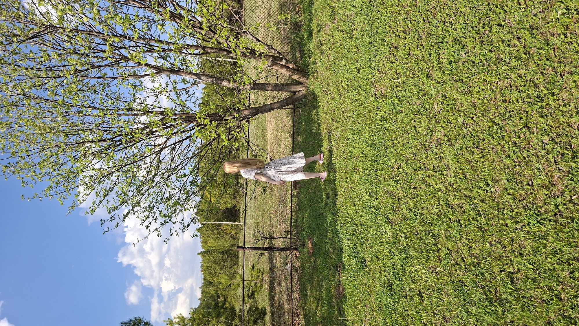 A young woman in a white dress stands in a grassy field in Большой Букор, Russia. A tree with green leaves is on the left. A fence separates the field from a wooded area. The sky is blue with white clouds.