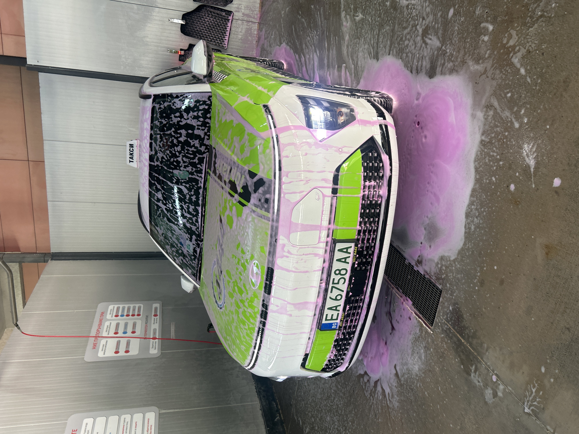 The image shows a white car with green accents being washed with pink soap. The car appears to be a taxi, as indicated by the sign in Ukrainian on the left side of the windshield that reads "ТАКСИ" (TAXI) and is at a car wash. The license plate, which is "EA 6758 AA" indicates that this car is located in Bulgaria.
The car is covered in pink soap suds, and there is also pink soap on the concrete floor around the car. In the background, there is a wall with car wash information posted.