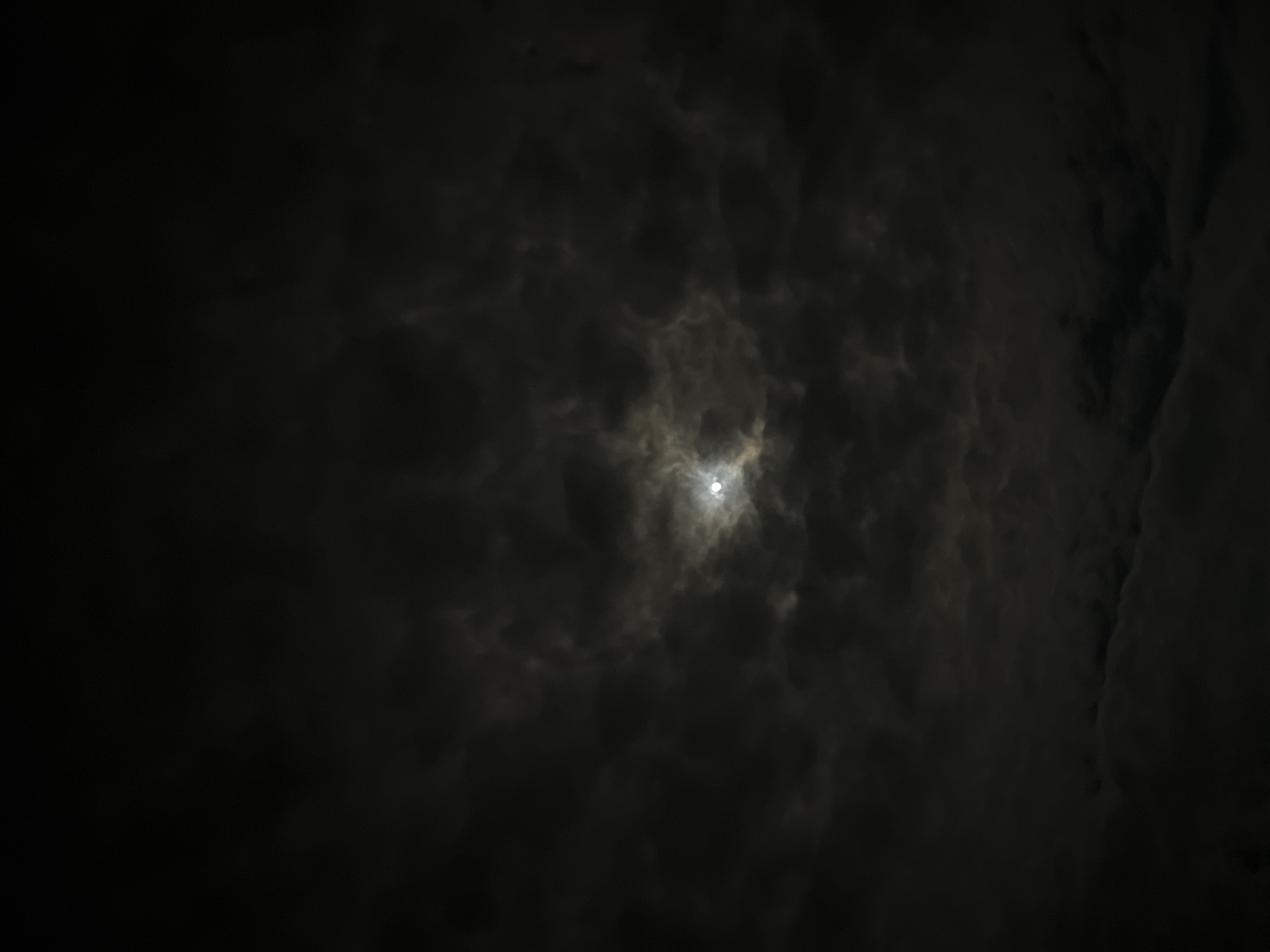 A night-time sky is depicted, filled with dark, textured clouds. A single, bright celestial object shines through a small break in the clouds, illuminating the surrounding cloudscape. The scene has a somber and mysterious atmosphere due to the heavy darkness and the obscured light source. There are no people, subjects, objects, actions, or interactions discernible in the scene. The photograph was supposedly taken in the city of Kotel, Bulgaria, but there is no discernible evidence of this visible in the image.