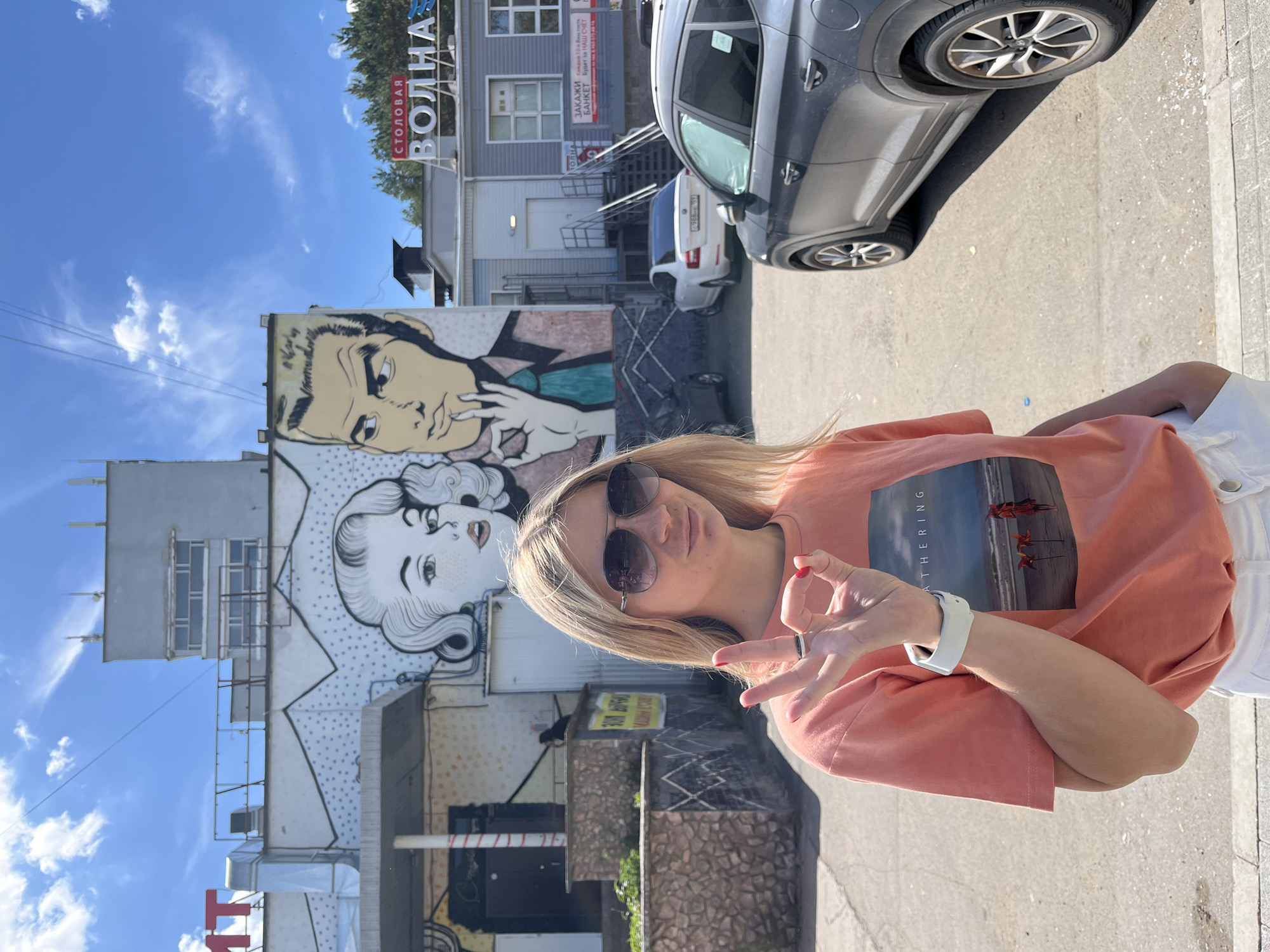 In Chaikovsky, Russia, a young woman with blonde hair and sunglasses stands in front of a building adorned with a pop-art mural of a couple. She is wearing a coral t-shirt with a graphic image and white shorts, and is holding up her right hand in an "okay" gesture, displaying a ring on her finger and red nail polish. A smartwatch is visible on her left wrist. The background features a blue sky with scattered clouds, a gray building with multiple windows, and several parked cars, including a dark gray SUV in the foreground. The setting appears to be an outdoor urban environment, possibly a street or parking lot.
