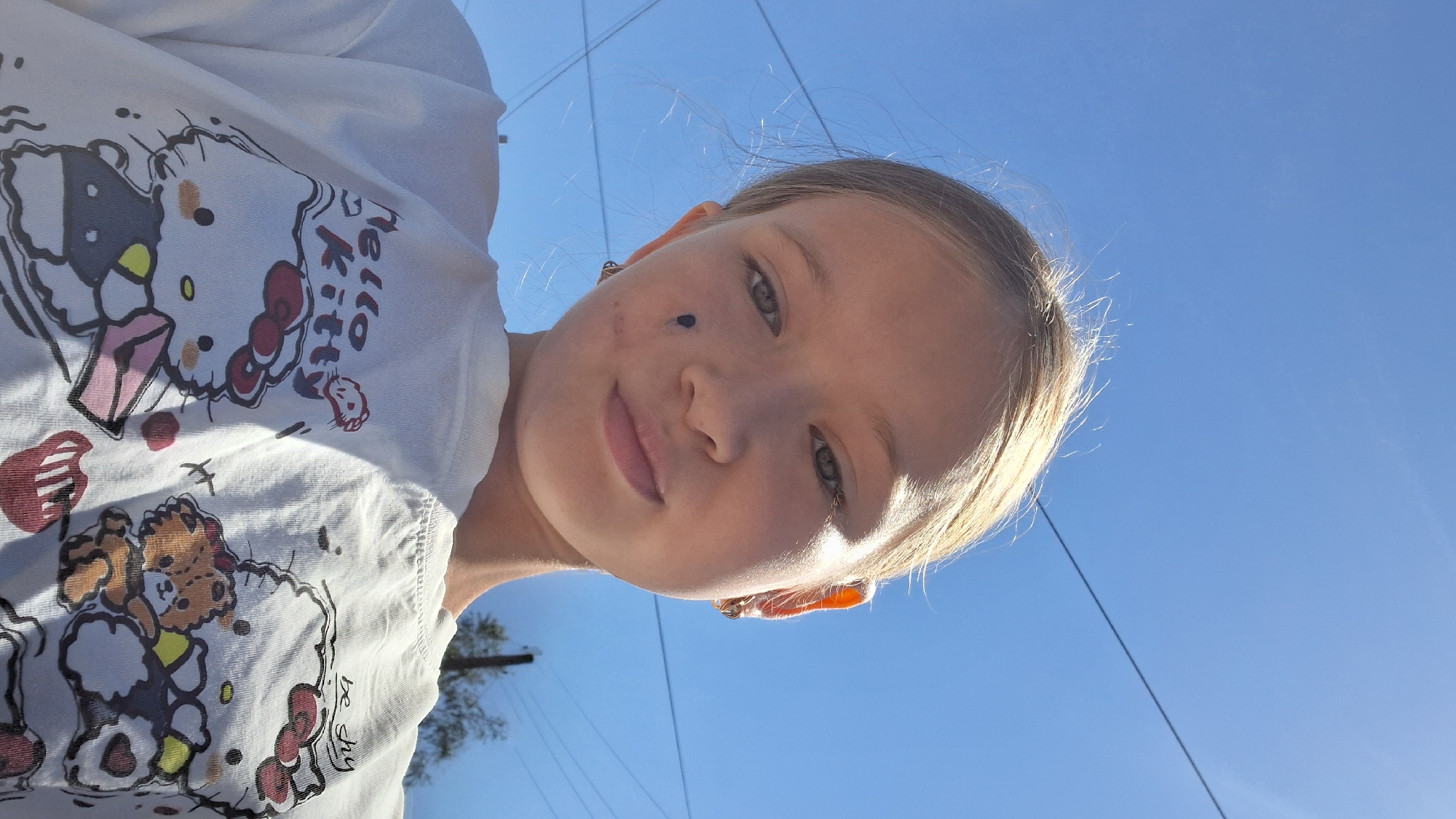 A young girl with blonde hair and a mole on her cheek is taking a selfie outdoors against a bright blue sky. She is wearing a white t-shirt with cartoon characters, including Hello Kitty and a teddy bear, printed on it. Her hair is windswept, suggesting it's a breezy day. The sunlight casts a warm glow on her face, and she has a slight smile. The image appears to have been taken during the daytime, given the bright sky. There are thin wires visible in the background, possibly power lines, and a glimpse of a tree. The overall mood is casual and cheerful. The t-shirt has some text, but it is difficult to read clearly in the image.