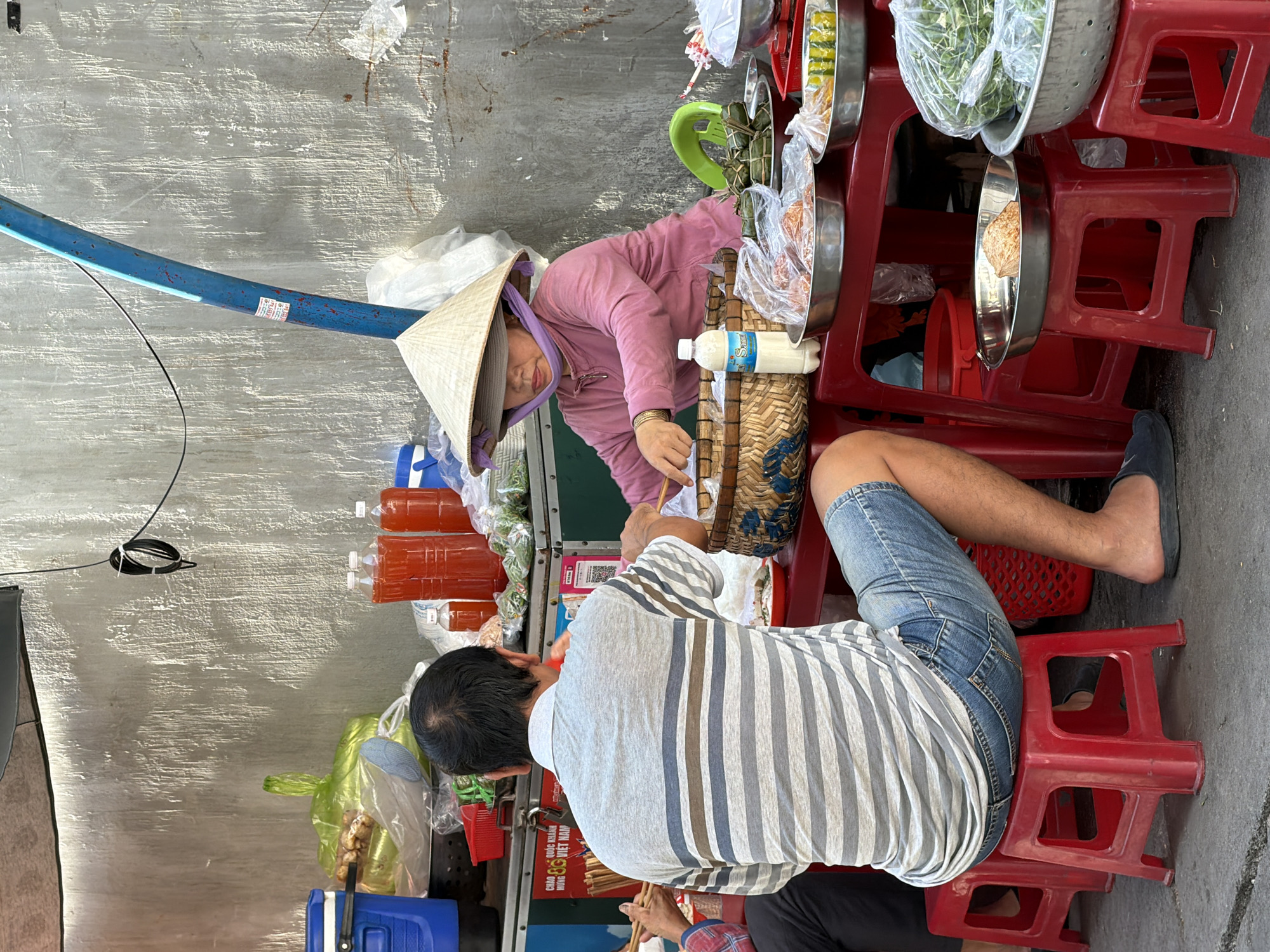 A street food vendor in Nha Trang, Vietnam, is serving a customer from a stall. The vendor, wearing a conical hat and a pink long-sleeved shirt, is seated behind a table laden with food. A man, the customer, sits on a red plastic stool facing the vendor. He is wearing a striped polo shirt and denim shorts. The customer is holding chopsticks and appears to be eating. The stall is organized with various food items displayed in metal bowls and plastic bags, including what appears to be leafy vegetables, prepared dishes wrapped in leaves, and bottles of red liquid, possibly drinks. The background shows a textured concrete wall and a blue metal pole, suggesting an outdoor setting, likely in a marketplace or along a street. The lighting indicates it is daytime.