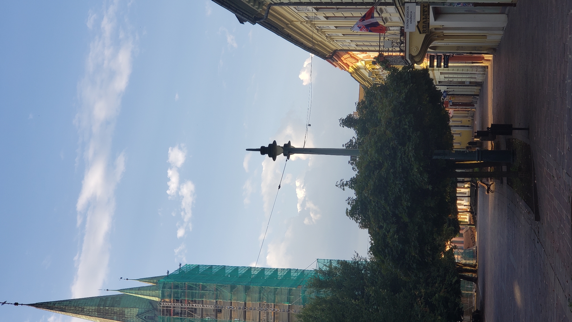 The image depicts an outdoor scene on a street in Prešov, Slovakia, likely during late afternoon or early evening, indicated by the long shadows cast by the buildings and trees. In the background to the left, a tall church spire, partially covered in green scaffolding, dominates the skyline. The spire is a prominent architectural feature, with smaller spires visible to its right. In the middle ground, a street lamp with a distinctive decorative top stands on a pole, with wires extending across the frame. Several mature trees with dense foliage are planted along the paved walkway, casting shadows onto the ground. Benches are visible beneath the trees, suggesting a public seating area. The right side of the image features a row of buildings with yellow and cream facades, exhibiting traditional architectural details and shop fronts. Two flags, one Slovakian, are visible on a balcony or window. The street is paved with interlocking bricks. The sky is blue with scattered white clouds.