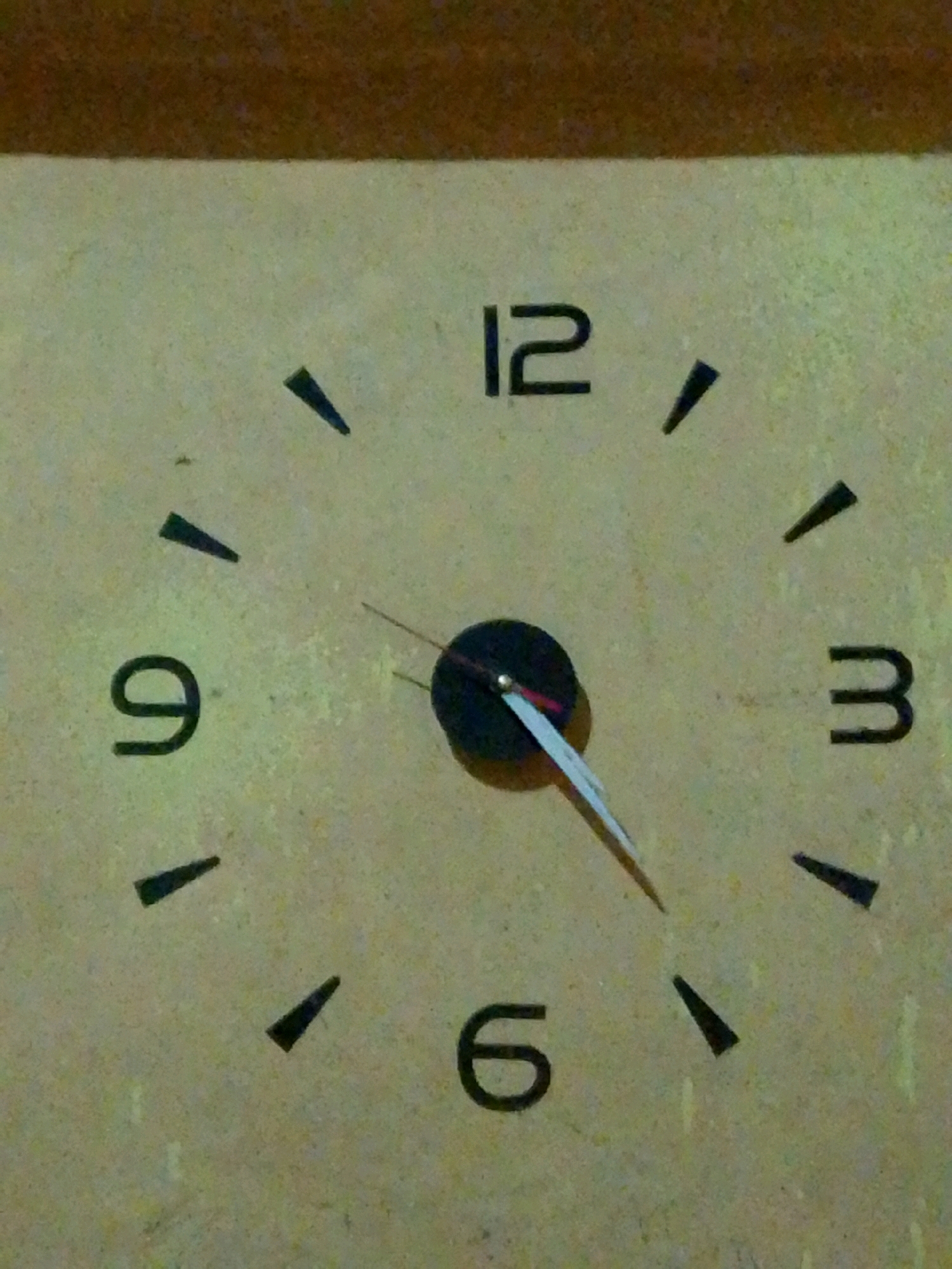 A close-up, head-on shot shows a clock face with black numerals and hour markers. The numerals 12, 3, 6, and 9 are clearly visible. The hour and minute hands are both pointing towards the 10 o'clock position, indicating a time of approximately 10:50. The clock face is light-colored and appears to be made of a textured material. The background is a blurred, dark brown. The image is well-lit, with the clock face being the primary focus. No people or other objects are visible in the frame.