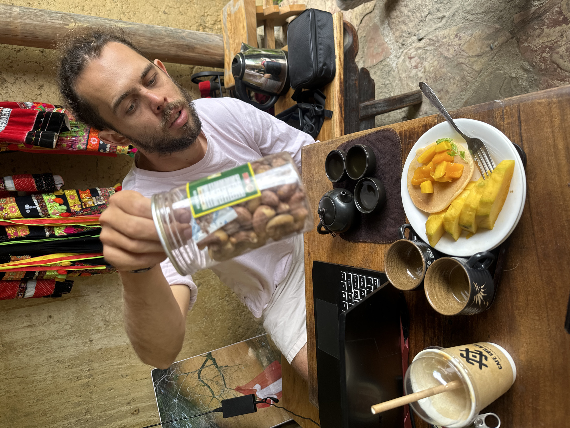 A man with brown hair and a beard is seated at a wooden table in Lũng Cú Commune, Vietnam. He is wearing a light pink t-shirt and white shorts. He is holding a clear jar filled with nuts.
On the table are:
* A laptop computer (black)
* A small teapot with four cups
* A white plate with a pancake topped with diced mango and slices of starfruit, along with a fork
* Two empty cups
* A paper cup with a clear lid and a straw.
In the background are:
* A shelf with colorful textiles
* A metal teapot and a black bag hanging on a wooden structure.
The walls are textured, possibly stone or stucco. A power adapter is visible below the table.