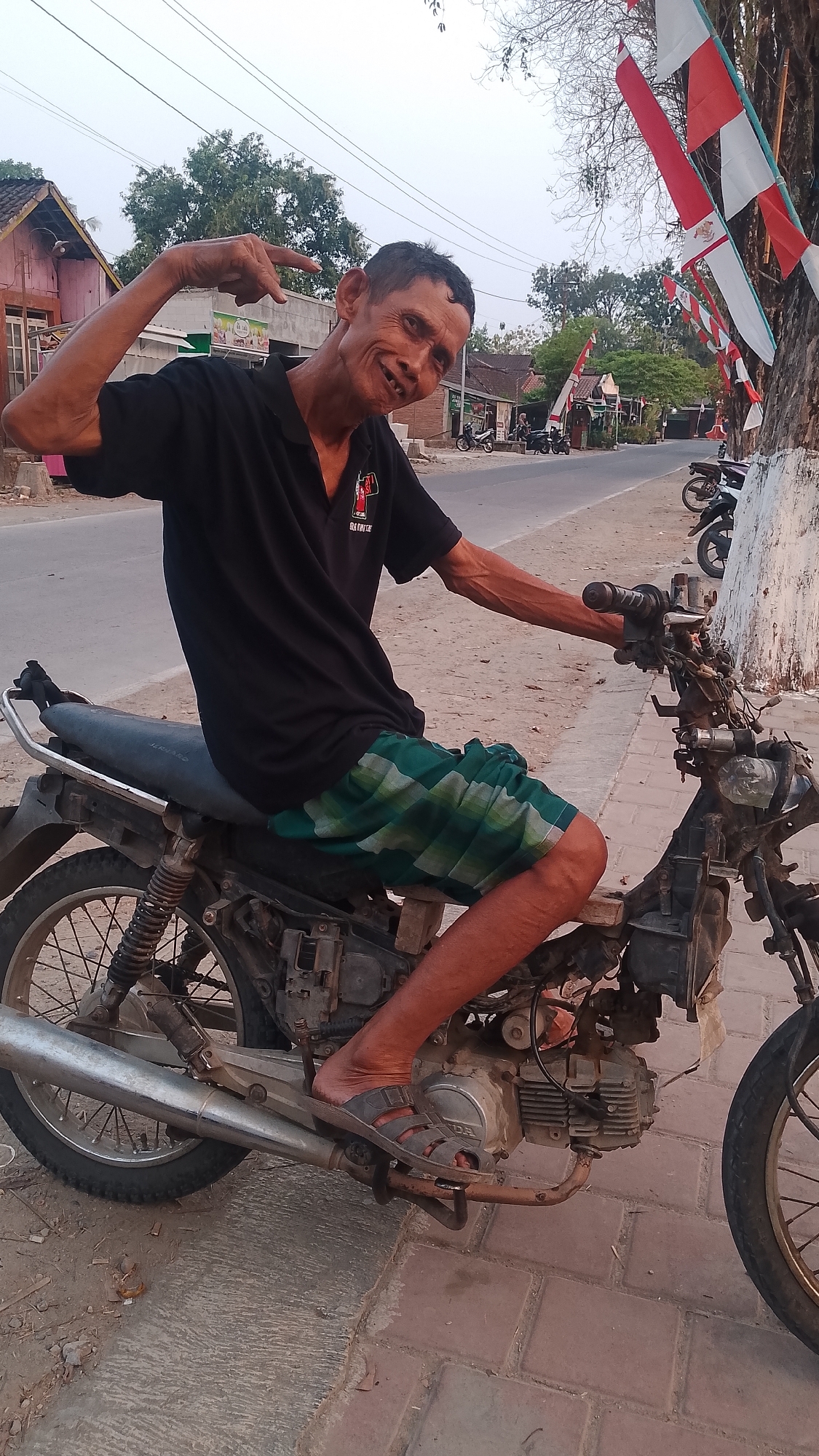 The image shows an elderly man sitting on a motorcycle on a road in Karanglangu, Indonesia. He is wearing a black polo shirt and a green and white checkered sarong. He is smiling and making a peace sign with his left hand. The motorcycle is old and appears to be in disrepair. There are buildings and trees in the background, as well as Indonesian flags hanging from a tree. The road is paved and there are other motorcycles parked in the background. The scene appears to be a street in a village or small town.