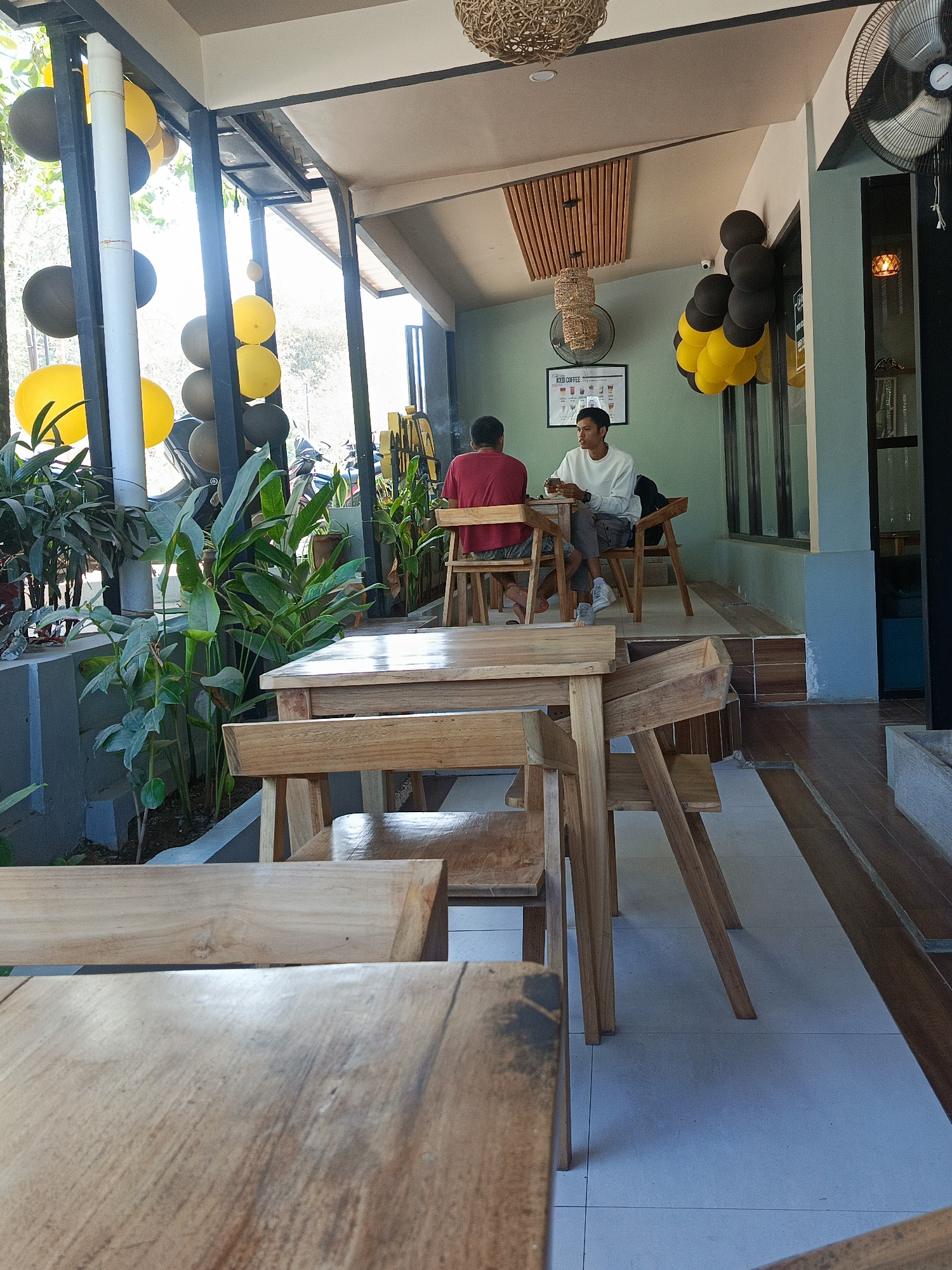 The image depicts the outdoor seating area of a cafe, likely located in Kuta, Indonesia. Two men are seated at a wooden table. One man, wearing a red t-shirt, is facing away from the camera, while the other, in a white long-sleeved shirt and grey pants, is facing the camera and holding a cup. The cafe features wooden tables and chairs, with lush green plants visible in the foreground. In the background, balloons in black and yellow are tied to a pillar and a wall, suggesting a festive occasion. A large industrial fan is mounted on the wall, and a ceiling fan is visible above. The flooring appears to be tiled. The overall ambiance is casual and relaxed, typical of a cafe setting in a tropical location.