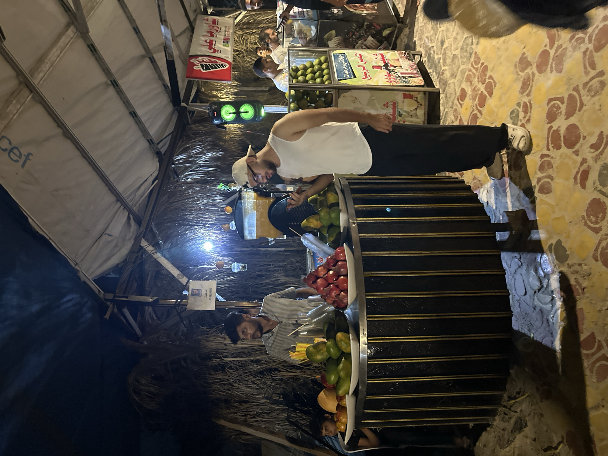 In Gaza City, an outdoor fruit juice stand operates at night under a covered structure, possibly made of tarpaulin and palm fronds. The stand displays a variety of fresh fruits, including mangoes, pomegranates, and oranges. Several individuals are present, with one man behind the counter tending to the fruits and another customer in the foreground observing the produce. The atmosphere suggests a local marketplace or street food vendor.