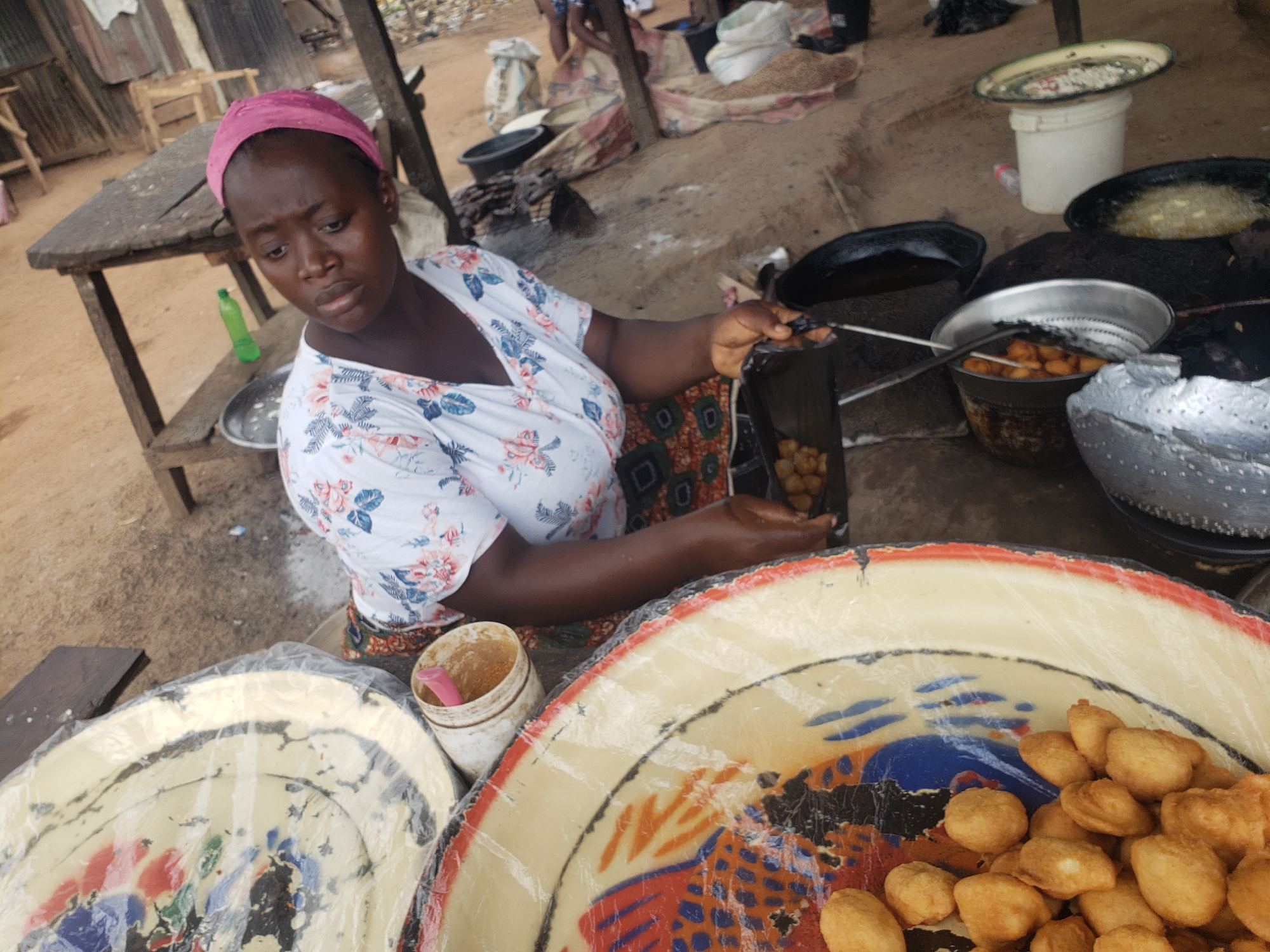 A woman, wearing a pink headscarf and a floral print shirt, is preparing food in an outdoor market stall, likely in Gumel, Nigeria. She is holding a black plastic bag containing fried dough balls and a metal slotted spoon. She is standing next to a large metal basin filled with more fried dough balls. In the background, there are several large metal pots, one containing bubbling oil, and other containers. A wooden table is to her left, and a green plastic bottle sits on a shelf beneath it. The ground appears to be dirt.