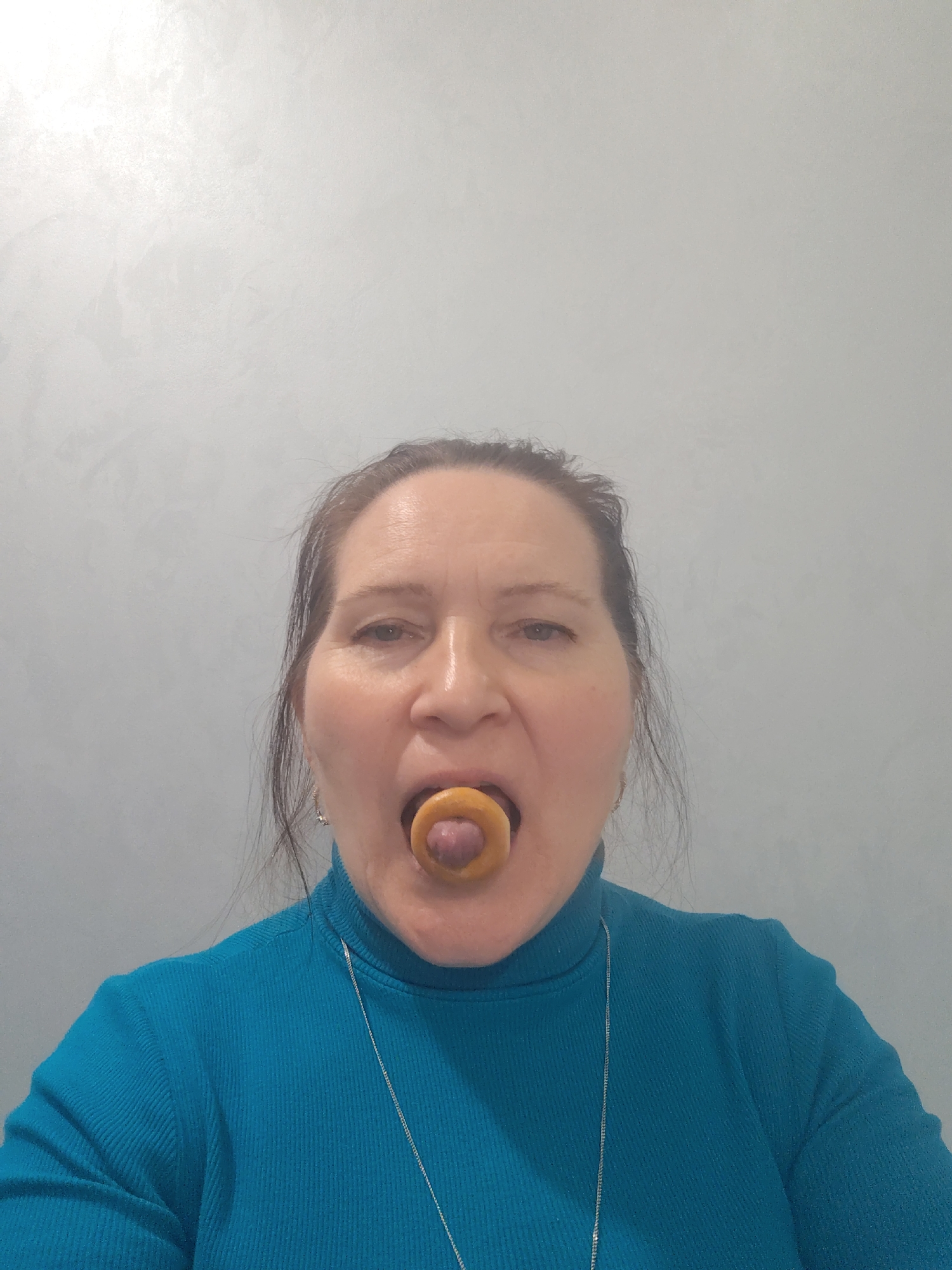 A middle-aged woman is depicted in a close-up, chest-up portrait against a light gray, textured wall. She has fair skin, dark hair pulled back, and is wearing a teal blue ribbed turtleneck sweater. A thin silver chain necklace is visible around her neck.
The central focus of the image is a small, light brown, ring-shaped pastry, possibly a bagel or a type of Russian "sushka" or "baranka," which the woman holds in her mouth. Her tongue is playfully extended through the hole of the pastry. Her eyes are open and directed towards the camera, and her expression is neutral, suggesting focus on the pose.
The background is a simple, uniformly lit wall with a subtle, uneven texture, possibly indicating a painted finish like venetian plaster. There are no other objects or details visible in the scene. The lighting appears even, suggesting an indoor setting. The overall impression is a lighthearted and candid moment. No text is visible in the image. The location context provided is Sarapul, Russia.