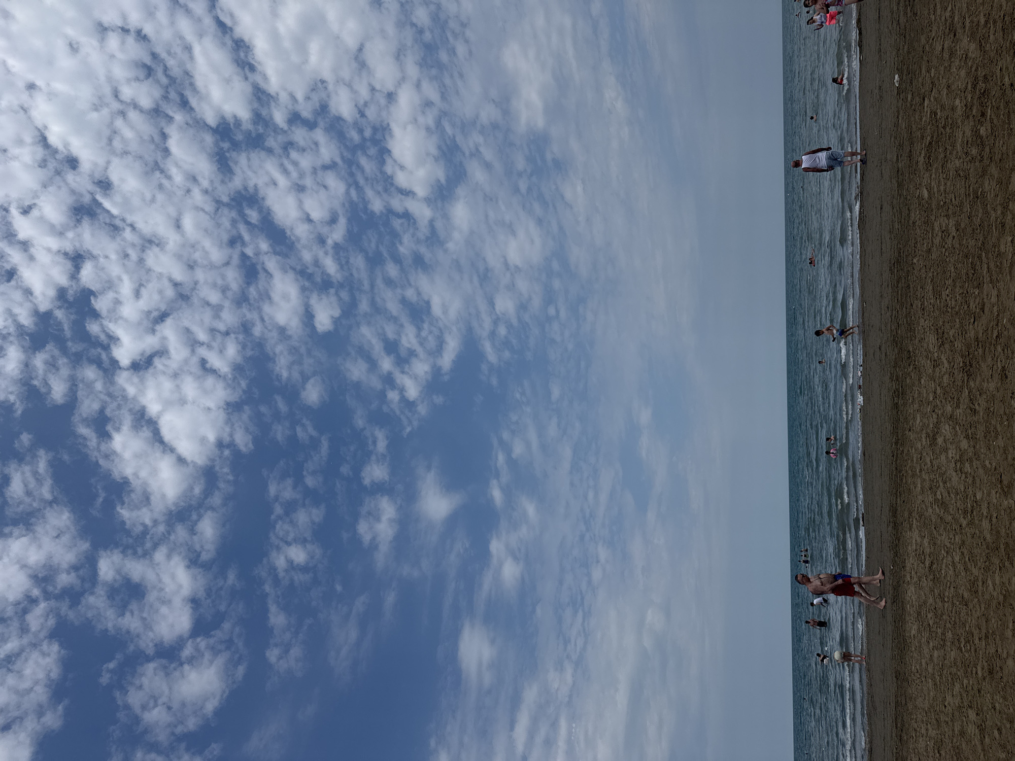 The image depicts a wide view of a beach in the city of Khazar Raion, Azerbaijan, under a sky filled with scattered white clouds. The foreground is dominated by a sandy beach, with the dark, wet sand closer to the water's edge. In the middle ground, a calm sea with gentle waves stretches to the horizon. Several individuals are visible on the beach and in the water. On the beach, a man in red shorts and another in blue shorts are walking from left to right. Further to the left, a person in red shorts and a white top is walking towards the sea. In the water, numerous people are swimming, wading, and playing. The water is a deep blue-green, and the sky above is a bright blue. The overall scene conveys a typical summer day at a recreational beach.