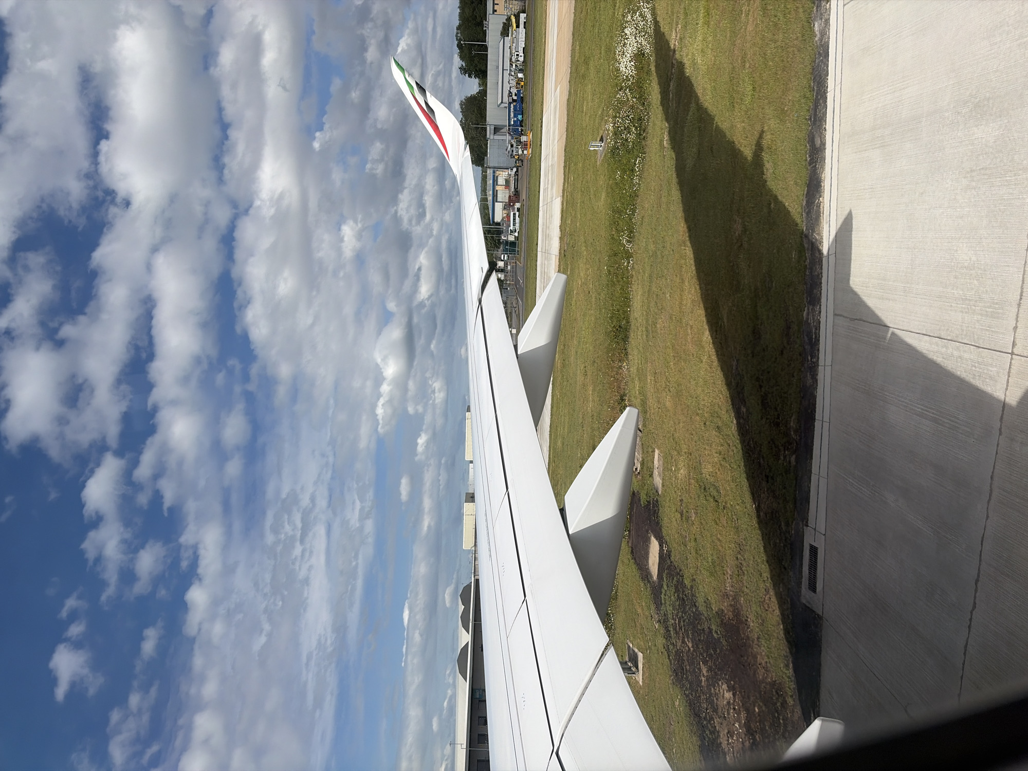 The image shows a view from inside an airplane, looking out over the wing and the airport grounds. The wing, painted white, takes up a large portion of the frame, and the Emirates logo is visible on the wingtip. The airport grounds below consist of a concrete surface and grassy areas. In the background, there are airport buildings and vehicles. The sky is visible with white clouds against a blue background. Based on the Emirates logo and the general appearance of the airport, the location is likely near Gatwick Airport, in the city of Crawley, United Kingdom.