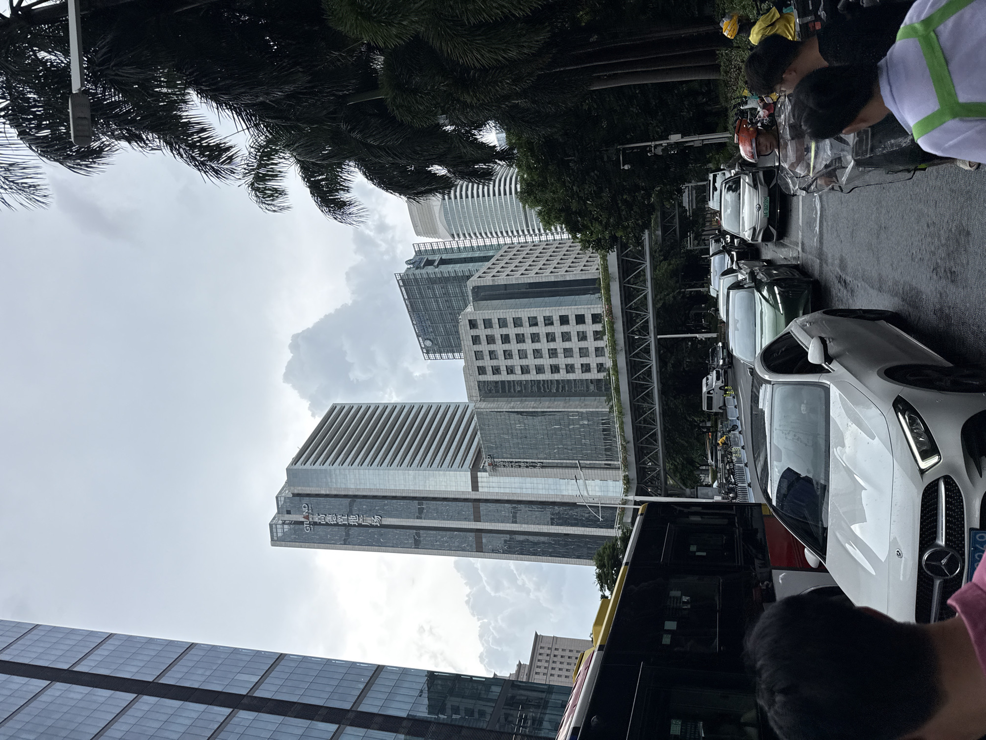 The image captures a street scene in Tianhe District, China, on a cloudy day, likely in the afternoon. The foreground is dominated by the side of a black bus and the front of a white Mercedes-Benz car. Several people are visible in the foreground, some of whom appear to be looking at something or engaged in conversation. Behind the vehicles, a line of traffic is visible, including other cars and motorcycles.
The background features a cluster of modern skyscrapers. One prominent building has a facade with horizontal lines, and another displays large text, partially obscured by a banner, that includes the characters "GT LAND" followed by Chinese characters. The sky is overcast, suggesting a possibility of rain or recent rainfall, as the road surface appears wet in places. Palm trees are present on the left side of the image, adding a tropical element to the urban landscape. The overall impression is one of a busy city street with considerable vehicle and pedestrian activity under a subdued sky.