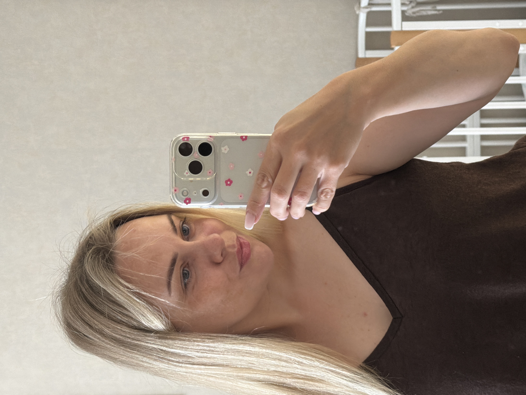 A young woman with blonde hair is taking a selfie in a mirror. She is wearing a dark brown v-neck top. She is holding a phone with a clear case decorated with small pink and white flowers. Her nails are painted a light pink. Her face is turned slightly towards the phone, and she has a slight smile. The background appears to be an indoor setting, possibly a bedroom, with a white crib or bed frame visible behind her. The lighting is soft and natural. The image does not provide any specific information to determine its location in the city of Votkinsk, Russia.