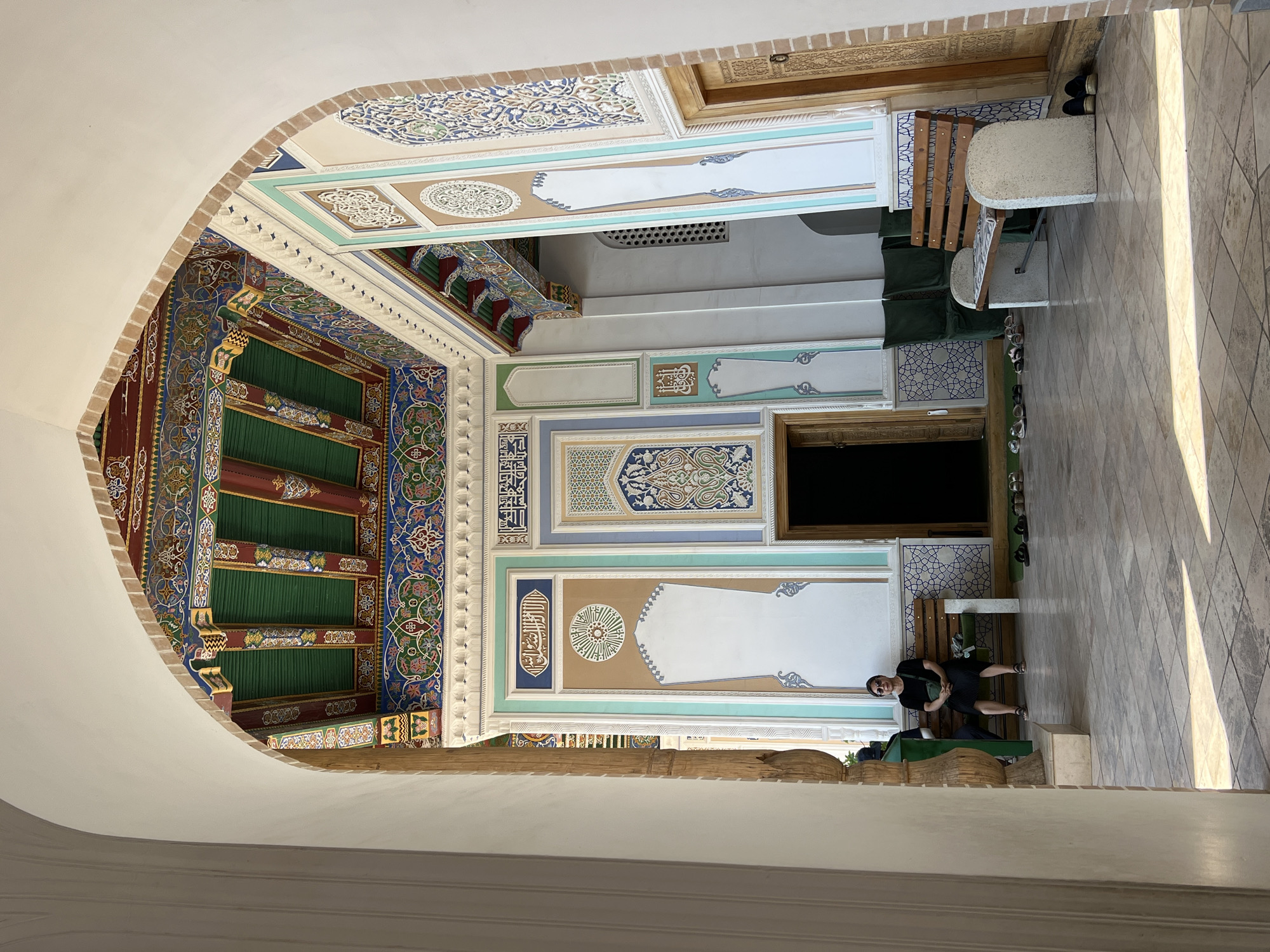 A person sits on a bench in a courtyard with ornate, colorful ceilings and walls. The architecture features intricate geometric patterns and Arabic calligraphy, indicative of Central Asian Islamic art. The ceiling is elaborately decorated with green, red, blue, and gold colors, featuring carved wooden beams. The walls are adorned with colorful tiled panels, some containing calligraphy. Sunlight streams into the courtyard, casting shadows on the tiled floor, suggesting it's daytime. The person, wearing sunglasses and a black outfit, appears relaxed. The setting looks like a historical site or a mosque courtyard in Samarkand, Uzbekistan, possibly a tourist area given the bench and the presence of shoes lined up near an entrance. No discernible weather conditions are apparent, but the light indicates a sunny day.