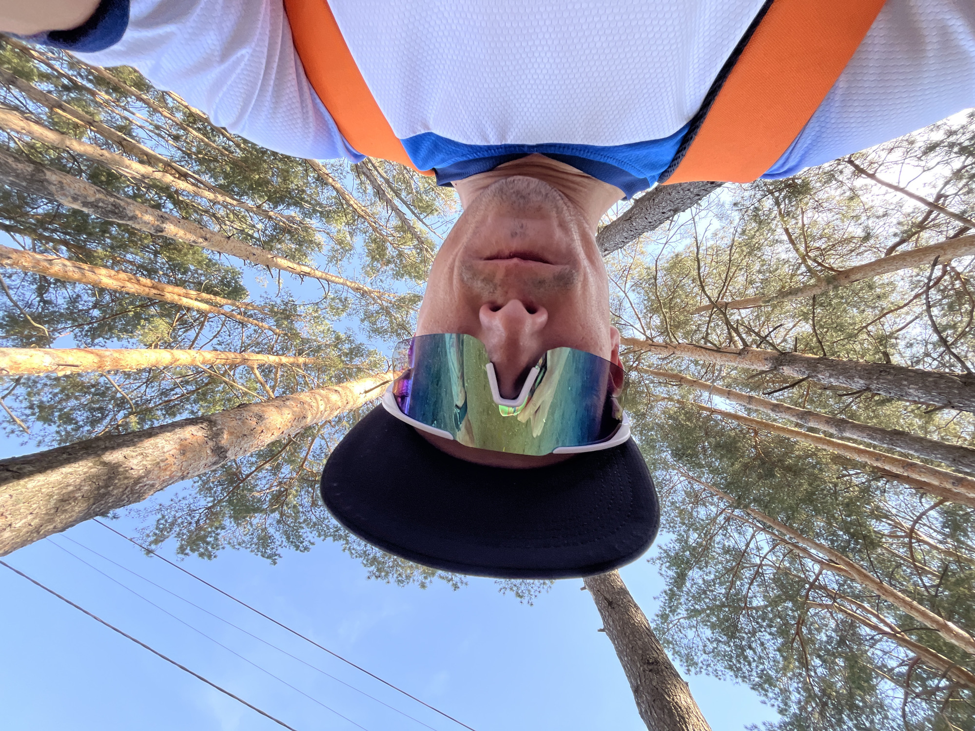 A man wearing sunglasses and a baseball cap is seen from an upside-down perspective. He has on a white, blue, and orange athletic shirt. Tall pine trees are visible in the background, reaching toward a bright blue sky. The photograph was taken in Chaikovsky, Russia, as the location was provided.