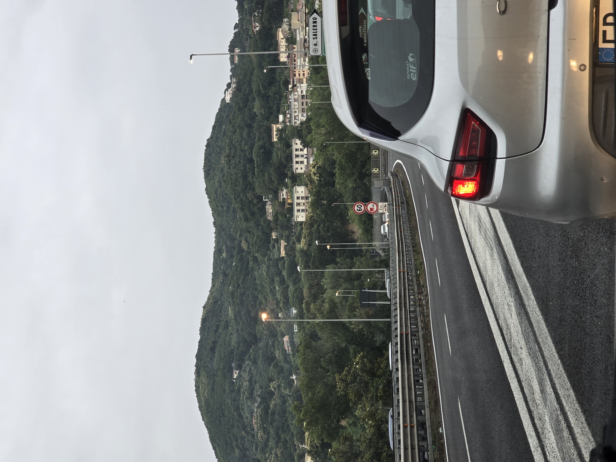 The image shows a view from inside a car, looking forward on a highway. The car's taillight is illuminated. In the background, a green, tree-covered hill rises with buildings scattered across its slope. A signpost on the right indicates "SALERNO" and "Amalfitana" with directional arrows, placing the scene in or near Salerno, Italy. The road ahead appears to be congested, with other vehicles visible. The sky is overcast.
