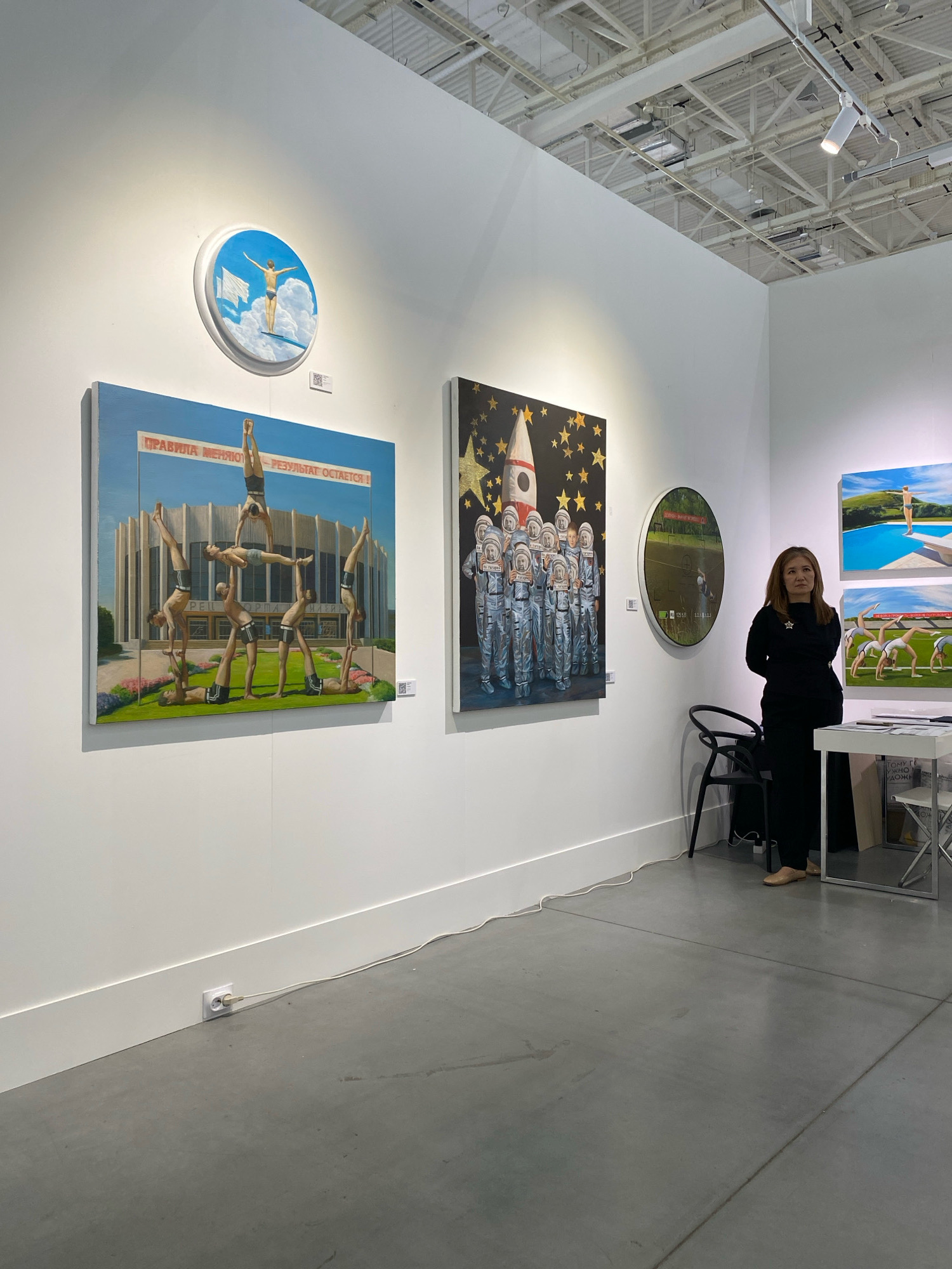 The image shows an indoor art exhibition at an unknown location, possibly in Khimki, Russia. The walls are white, and the floor is a polished concrete. Several artworks are displayed, including a circular painting of a diver on a cloud, a rectangular painting of a human pyramid in front of a modern building, and a painting of astronauts with a rocket. There is a woman standing near a table with art books. The exhibition space appears well-lit with track lighting.