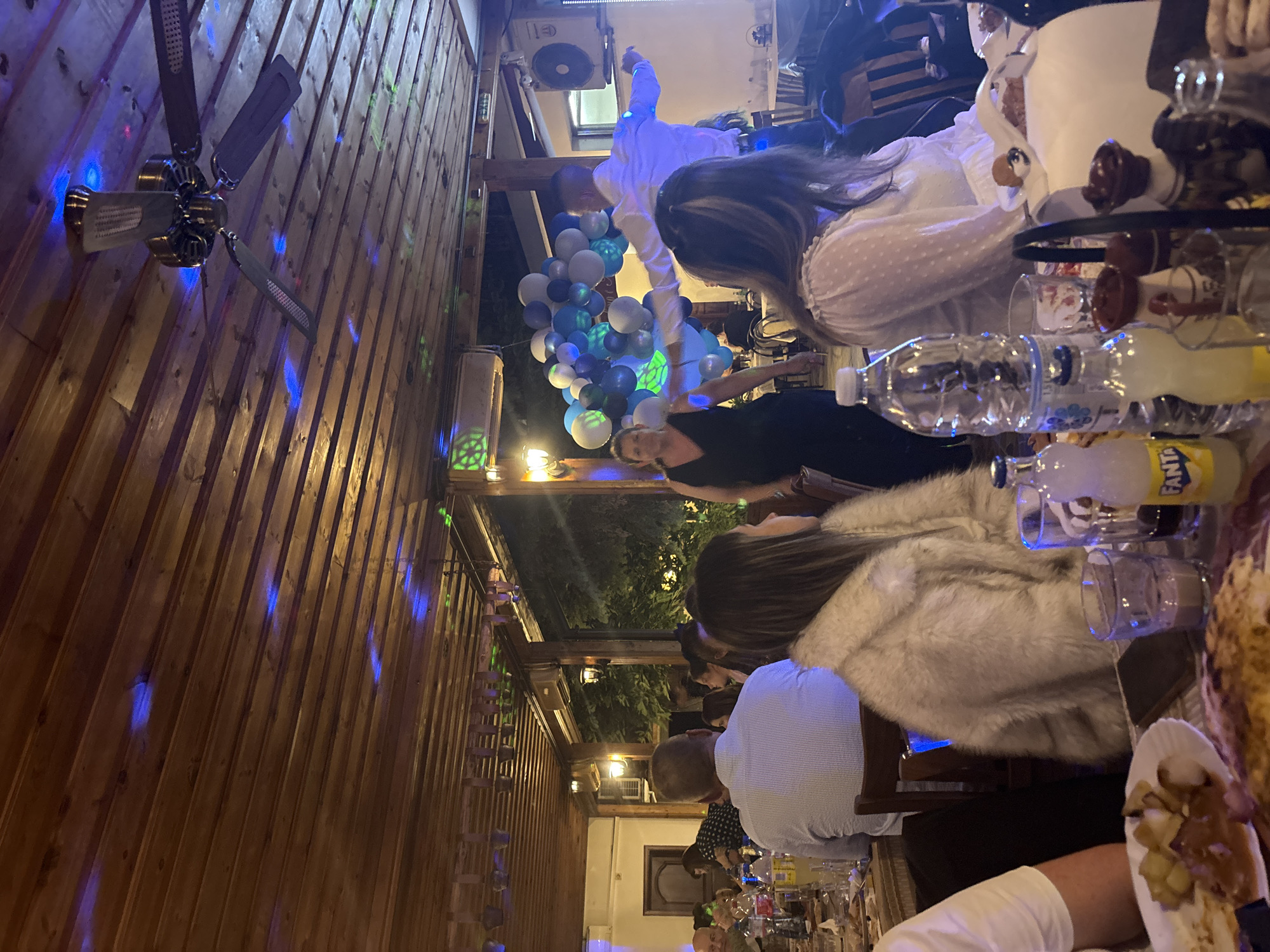 The image depicts an indoor gathering, possibly a celebration, at a restaurant in Karlovo, Bulgaria. The scene is dimly lit, with a wooden ceiling and exposed wooden beams above. Several people are seated at tables, engaged in conversation and dining. In the background, a man in a white shirt is dancing, with a backdrop of blue and white balloons. A woman in a black dress stands to his left, observing. There are bottles of beverages on the tables, along with plates of food. A ceiling fan is visible, and colored lights, primarily blue, are projected onto the ceiling, creating a festive atmosphere. The overall impression is one of a lively social event.