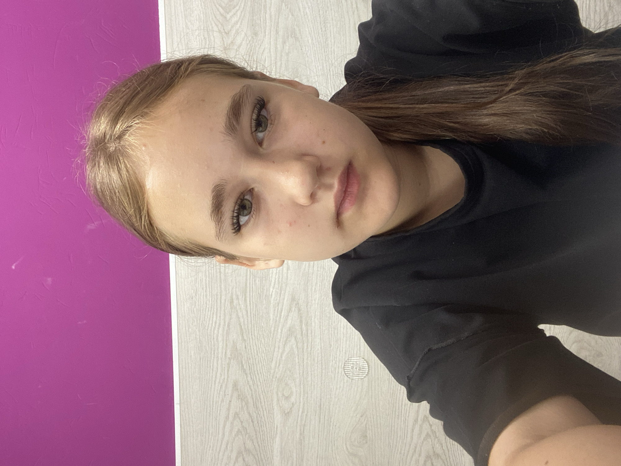 A young woman with light brown hair pulled into a ponytail is looking directly at the camera. She is wearing a black t-shirt. She has green eyes and is wearing false eyelashes. The background consists of a bright pink wall and a light wood-paneled wall. There is a small, circular, patterned object on the wood-paneled wall to the left of the woman. The lighting appears to be from overhead. The image is a selfie taken at eye level. The woman is positioned in the center of the frame. The overall impression is casual and personal. There are no discernible elements that would indicate the specific location in the city of Vysoki, Russia.