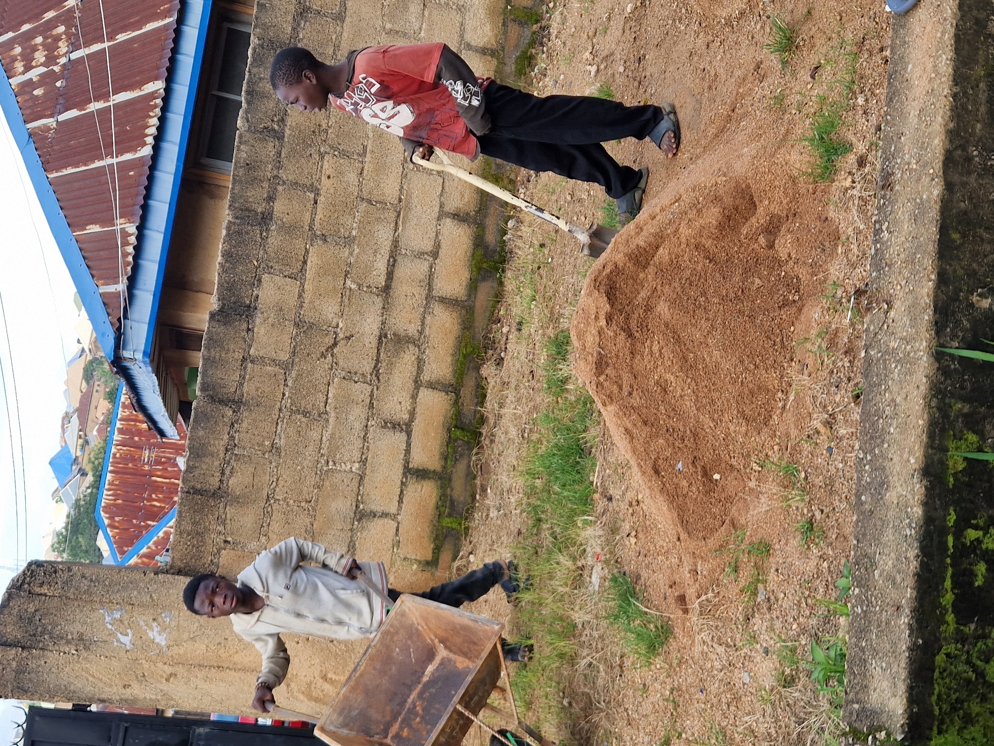Two young boys are engaged in manual labor near a pile of sand in Kabong, Nigeria. The boy on the left, wearing a light-colored hooded jacket and dark pants, is pushing a rusty wheelbarrow. The boy on the right, dressed in a red t-shirt with a graphic design and black pants, holds a shovel, appearing to be working with the sand. They are positioned on a dirt and gravel ground with sparse vegetation. In the background, a brick wall separates the work area from a residential area with buildings featuring corrugated metal roofs, suggesting an informal urban or semi-urban setting. The sky is overcast.