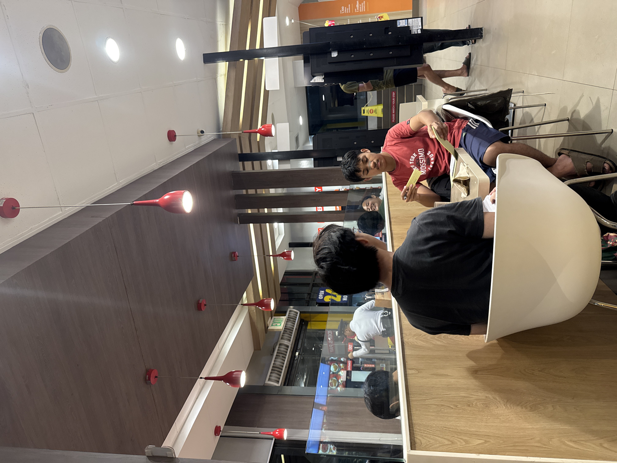 This image, presented with a tilted perspective, captures an indoor scene within a casual restaurant or food establishment in Davao City, Philippines, likely during the evening or night due to the reliance on artificial lighting.
In the foreground, a person with dark hair, wearing a black t-shirt, is seated in a sleek white chair, facing away from the camera. To their right, another young man with short dark hair, clad in a red t-shirt with "NEW YORK MOSSIMO" printed on the front, is seated. He is actively engaged with a light yellow or green item, possibly a food wrapper or a small menu, held in his hands. He wears dark shorts and brown slip-on sandals. Visible in the background are the legs and feet of other patrons, some also wearing sandals.
The restaurant's interior features a modern design with a white ceiling adorned with circular recessed lights and a speaker, complemented by several distinctive red pendant lights hanging on thin wires. Walls and partitions are covered in wooden-textured panels, contrasting with prominent black structural elements. Glass partitions create reflections, showing parts of the interior and what appears to be an outdoor area.
Visible text includes "NEW YORK MOSSIMO" on the red t-shirt, a green "EXIT" sign, and a yellow sign on the floor stating "PLEASE WATCH YOUR STEP." Reflections in the glass also show partial text like "OPEN" and "YAG & BRO." The scene depicts a relaxed, casual dining environment where people are seated and engaging with their food or company.