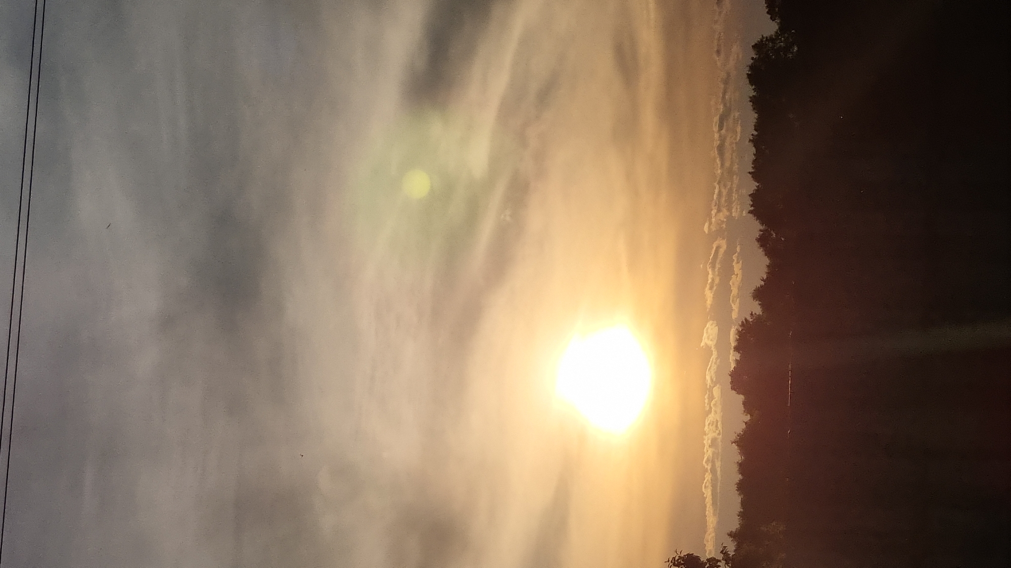 The image shows a sunset over a treeline. The sun is a bright, glowing orb in the sky, casting a warm, golden light. Wispy clouds are scattered across the sky, tinged with pink and orange. The treeline is silhouetted against the sky, with dark, dense foliage. There are power lines visible at the top of the image. The foreground is dark, suggesting it is a field or open area. The scene is serene and peaceful, with the natural beauty of the sunset dominating the frame. The location is identified as Vysokoe, Russia.