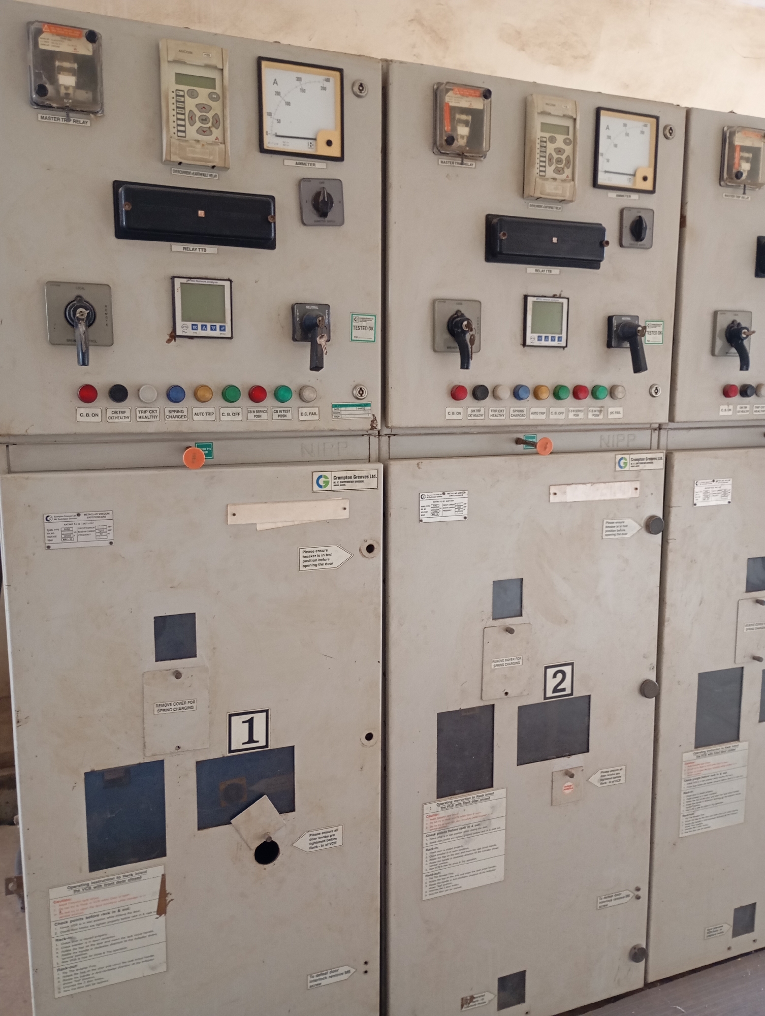 The image displays a close-up view of an electrical control panel, likely situated in an industrial or utility setting. The panel is gray and appears to be made of metal. It is divided into multiple sections, each containing various instruments and controls.
On the upper portion of the panel, there are several rectangular and square devices. These include what appear to be trip relays, an ammeter with a dial displaying current in amperes, a digital display unit, and multiple switches or buttons. Labels are present beneath some of these devices, such as "MASTER TRIP RELAY," "AMMETER," and "RELAY TTb." There are also rows of colored indicator lights (red, yellow, green, blue, white) with labels like "C.B. ON," "TRIP CKT HEALTHY," "SPRING CHARGED," "AUTO TRIP," "C.B. OFF," "CB IN SERVICE," "CB IN TEST POSN," and "D.C. FAIL."
Below the upper section, there are larger cabinet-like compartments. One compartment on the left has a prominent label with the number "1" in a black square. Another compartment to its right has a label with the number "2." These compartments have doors with circular knobs and some have warning labels or instructions. For instance, one label on the compartment with "1" reads, "Operating Instructions to Rack In/out the VCB with front door closed." Another on the compartment with "2" appears to have similar operational instructions. There are also company logos visible, such as "Crompton Greaves Ltd."
The overall environment appears to be indoors, with a plain, neutral-colored wall visible in the background. The lighting is somewhat subdued. The presence of the electrical equipment suggests a functional facility related to power distribution or control. The image does not contain any people.
The prompt mentions the city of Shendam, Nigeria. While the image itself does not contain any direct visual cues to pinpoint its exact location within Shendam, the presence of the electrical panel in an industrial or utility context is consistent with infrastructure found in such a city.