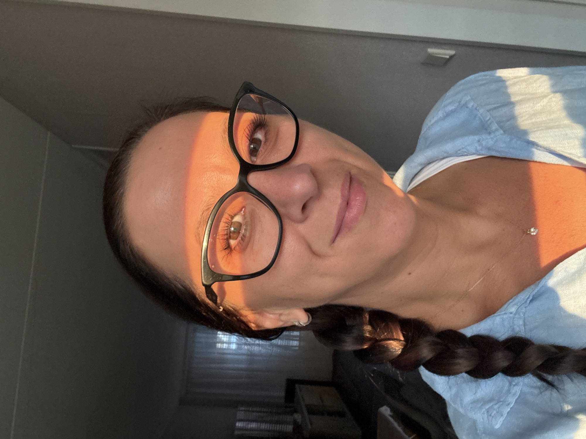 A medium shot shows a woman wearing black-rimmed glasses and a light blue collared shirt. Her dark hair is braided and hangs over her left shoulder. A strand of sunlight illuminates the left side of her face and forehead. She is wearing a small necklace with a diamond pendant. The background is dimly lit, with hints of furniture and a window with blinds visible on the left side of the frame. The right side of the frame shows a grey wall. This photograph was taken in Marum, Netherlands.