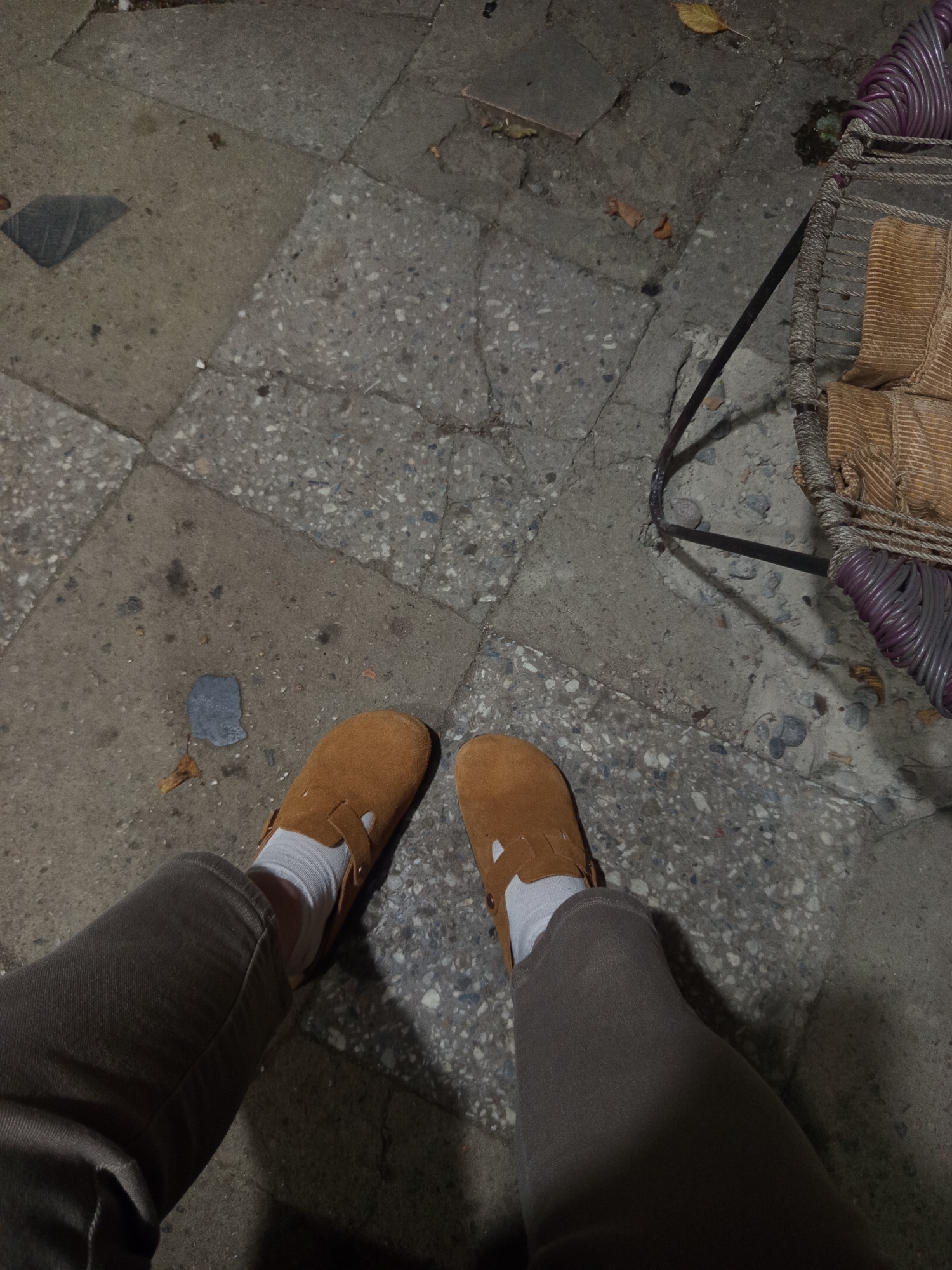 The image captures a ground-level view of a person's legs and feet, clad in brown suede slip-on shoes and white socks, standing on a paved surface composed of large, irregular stone tiles. The tiles exhibit variations in color and texture, with some appearing lighter and speckled, while others are darker and smoother. Scattered across the pavement are small leaves and debris. To the right of the frame, a portion of a woven chair is visible, featuring a dark metal frame and a seat with light brown fabric. The lighting suggests it is either evening or a dimly lit area, casting subtle shadows on the ground. The scene is set in the city of Khashuri, Georgia.