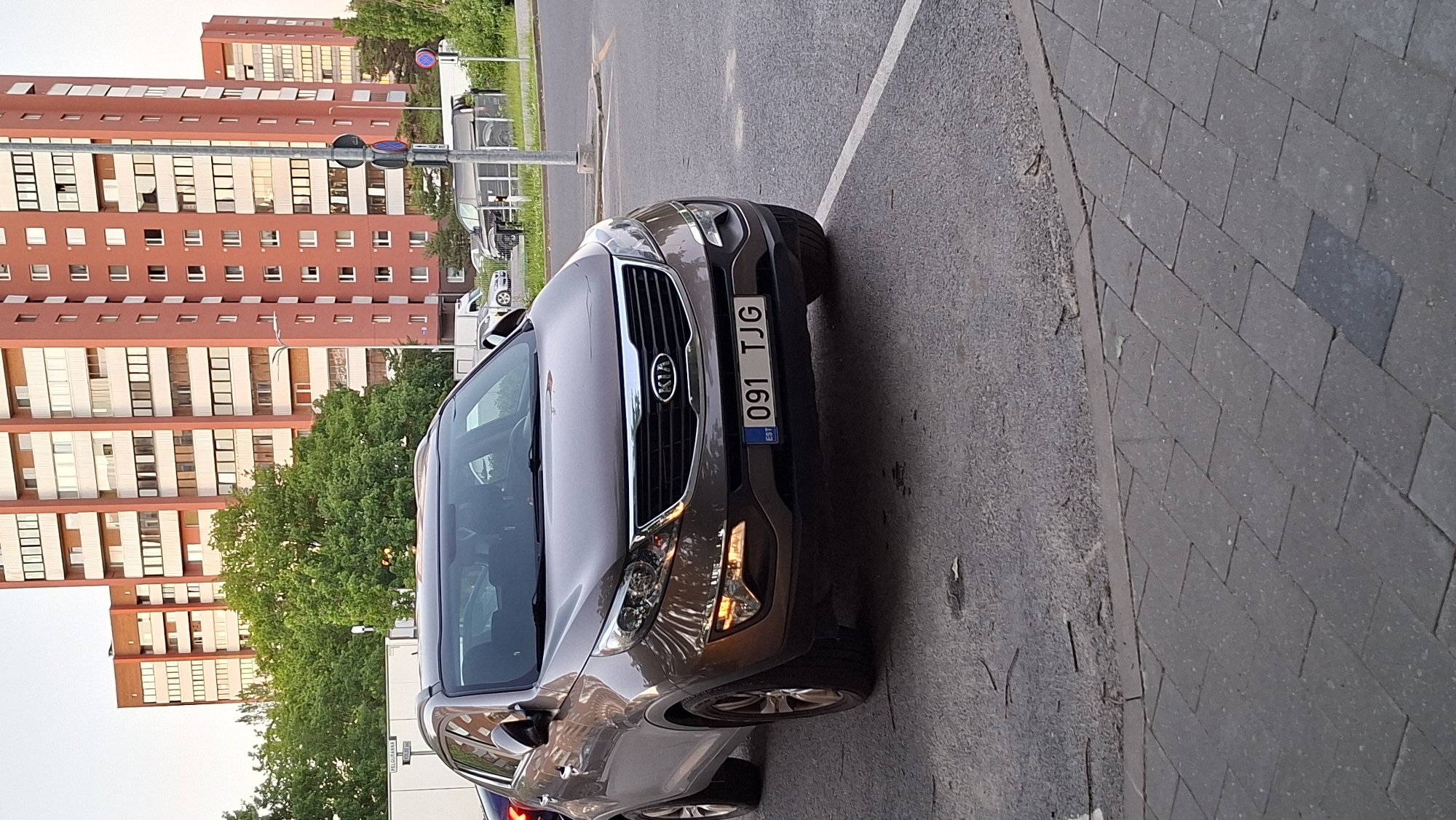 A brown Kia Sportage is parked on a paved area next to a sidewalk. The car has a license plate that reads "091 TJG EST". In the background, there are tall brick buildings with many windows, green trees, and street signs. The scene appears to be in an urban environment, likely a parking lot or street in Tallinn, Estonia. There are no people visible in the image.