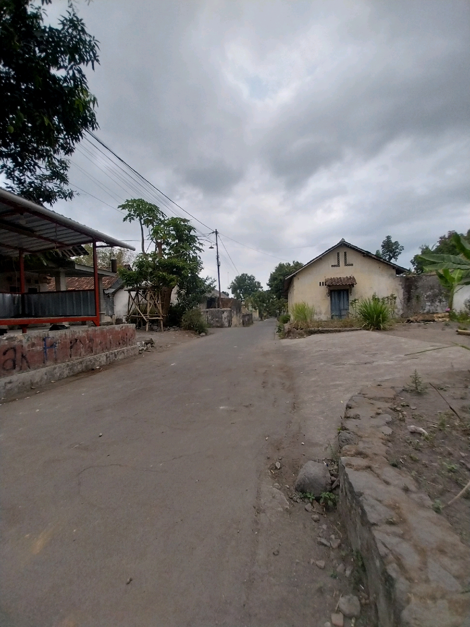The image depicts a rural street scene in Tijayan, Indonesia, under an overcast sky. A paved road runs through the center of the frame, curving slightly to the right. On the left side of the road, there is a covered structure with a corrugated metal roof supported by red metal posts, and a low brick wall with graffiti. Behind this structure are glimpses of buildings with tiled roofs. Further down the road, a large tree with scaffolding around its base stands near some smaller buildings.
On the right side of the road, a single-story house with a light-colored facade and a dark blue door is visible. The house has a tiled roof and decorative elements above the windows. The area around the house is somewhat overgrown with grass and vegetation. A stone wall borders the property on the right. The overall environment appears to be a modest residential area in a village setting. There are no people or animals visible in the image.